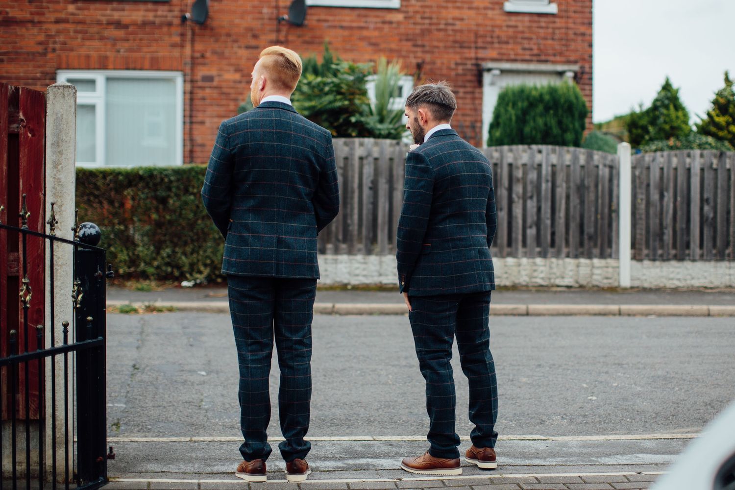 Two people in suits standing on a driveway in front of brick houses.