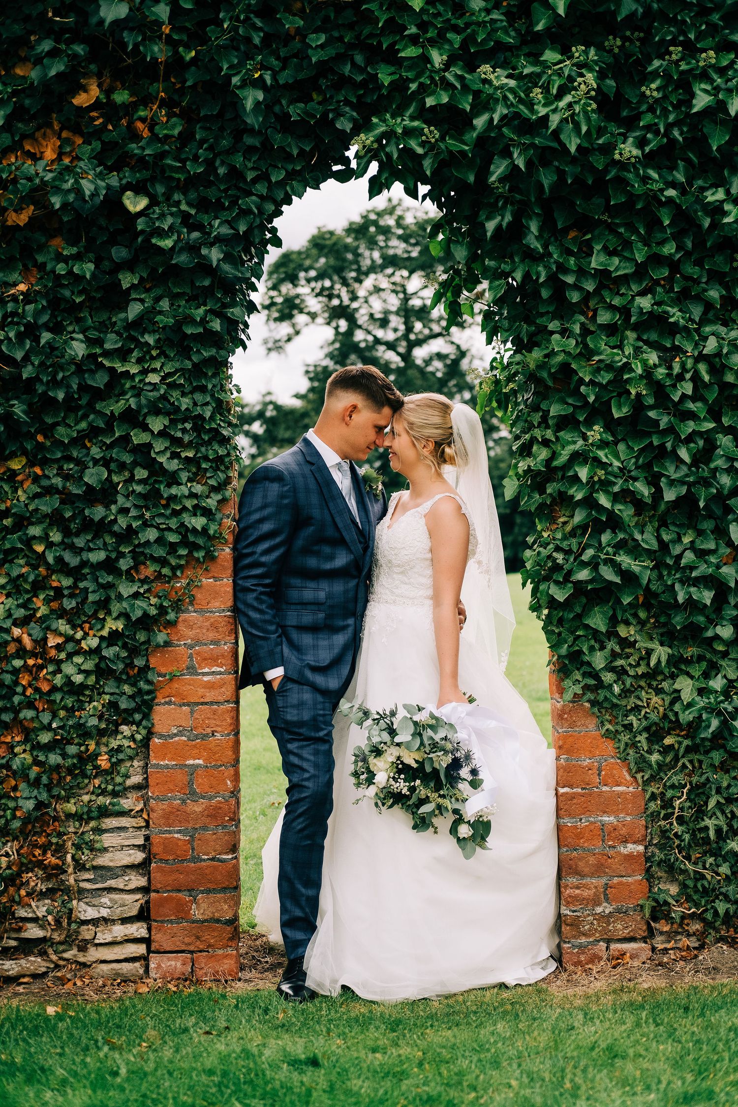 Newlyweds pose against ivy-covered brick wall in romantic garden setting.