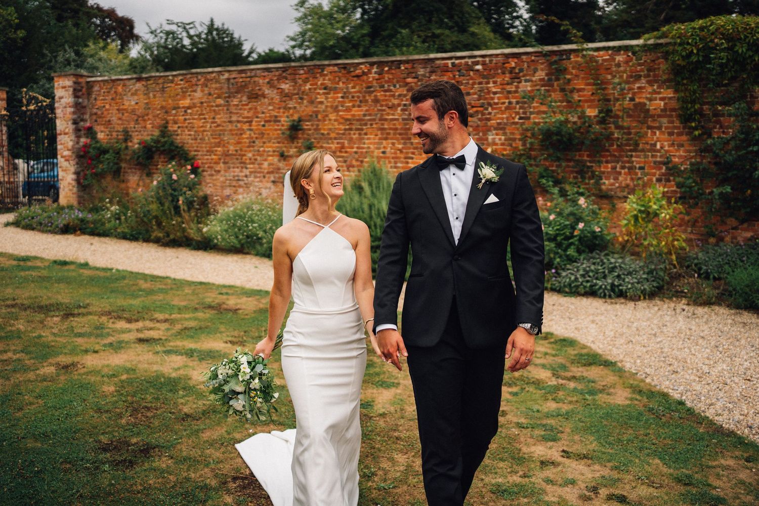 Newlywed couple walks together along garden path next to rustic brick wall on wedding day.