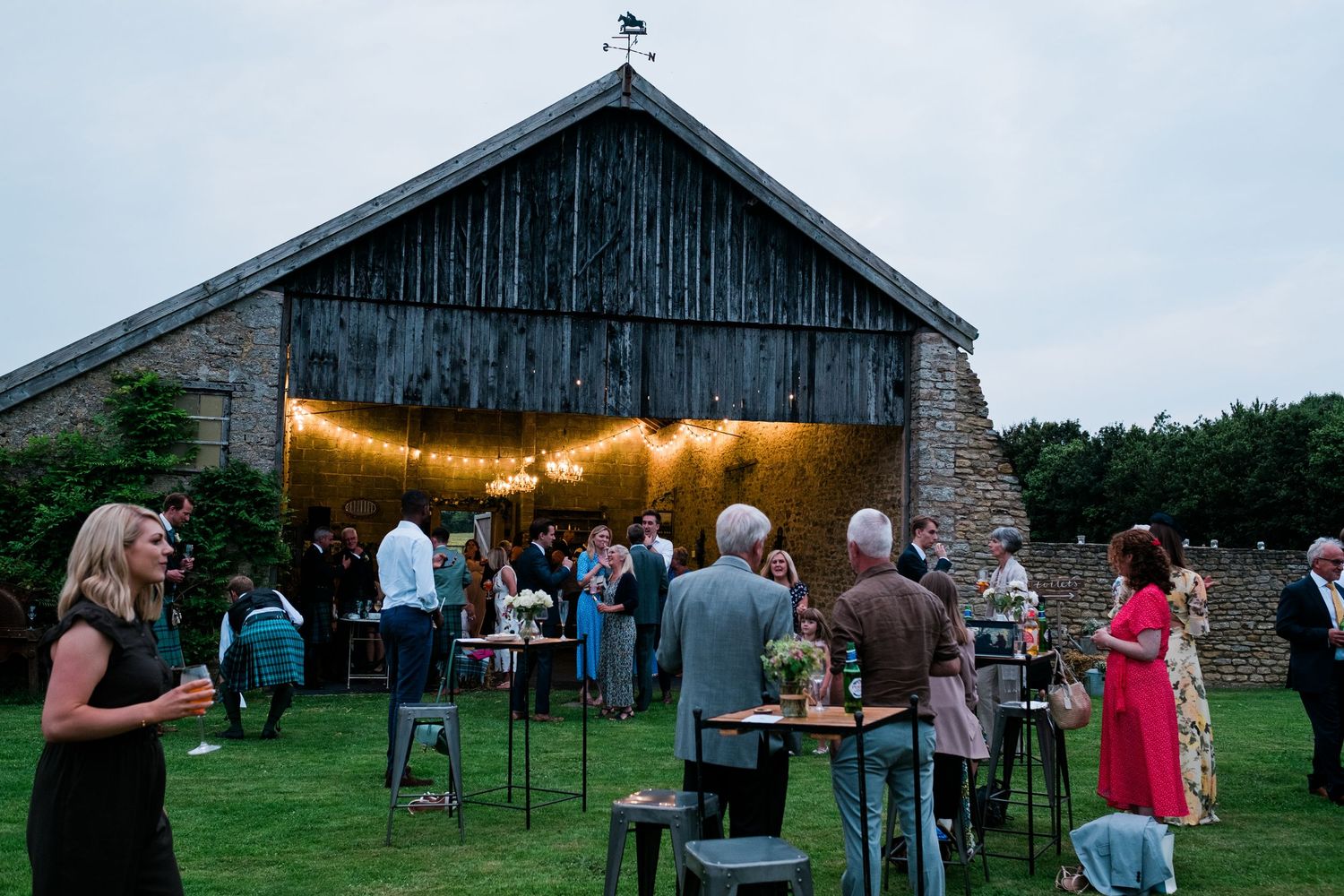 A rustic barn wedding venue at dusk with string lights and guests mingling on the lawn.