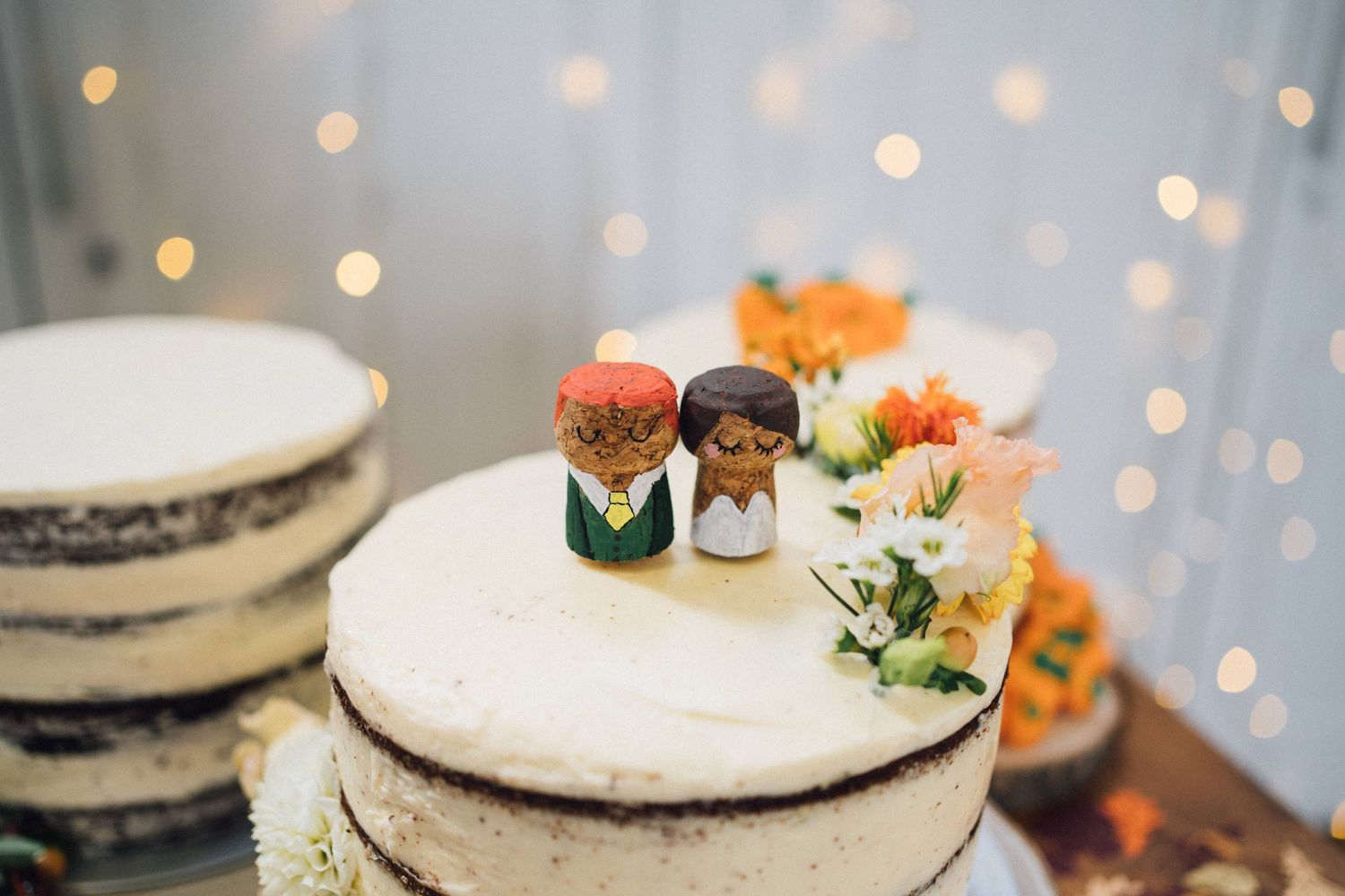 Rustic wedding cake decorated with fresh flowers and succulents on a wooden table with twinkling lights in background.