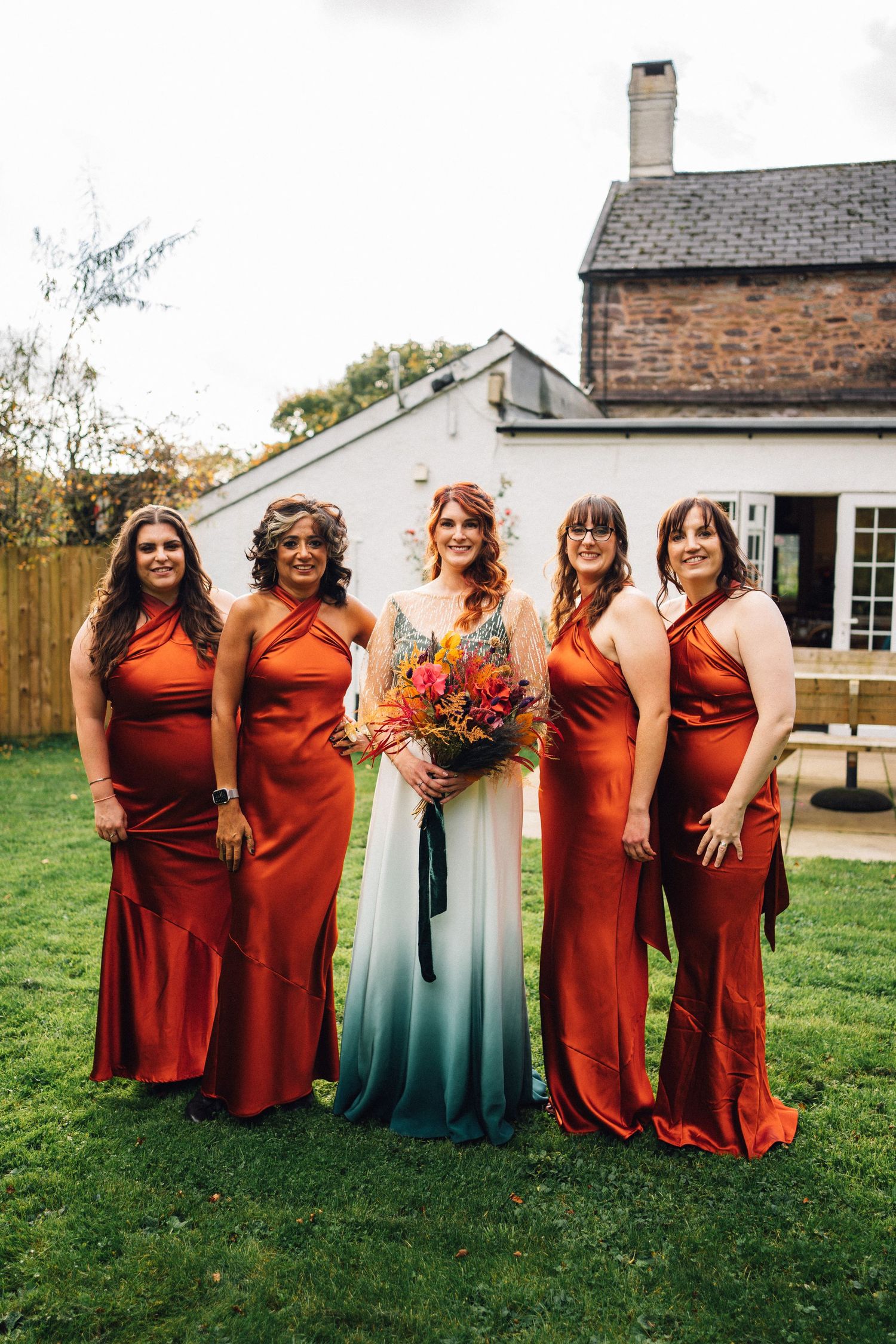 Wedding party poses on lawn wearing burnt orange bridesmaid dresses with bride in white and blue ombre gown.