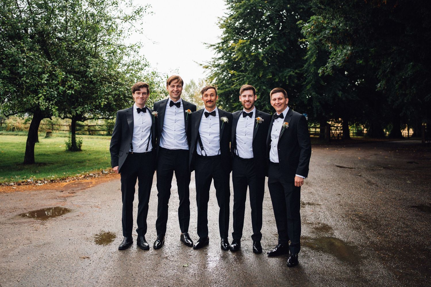 Groomsmen in black tuxedos walking together outdoors on a tree-lined path.