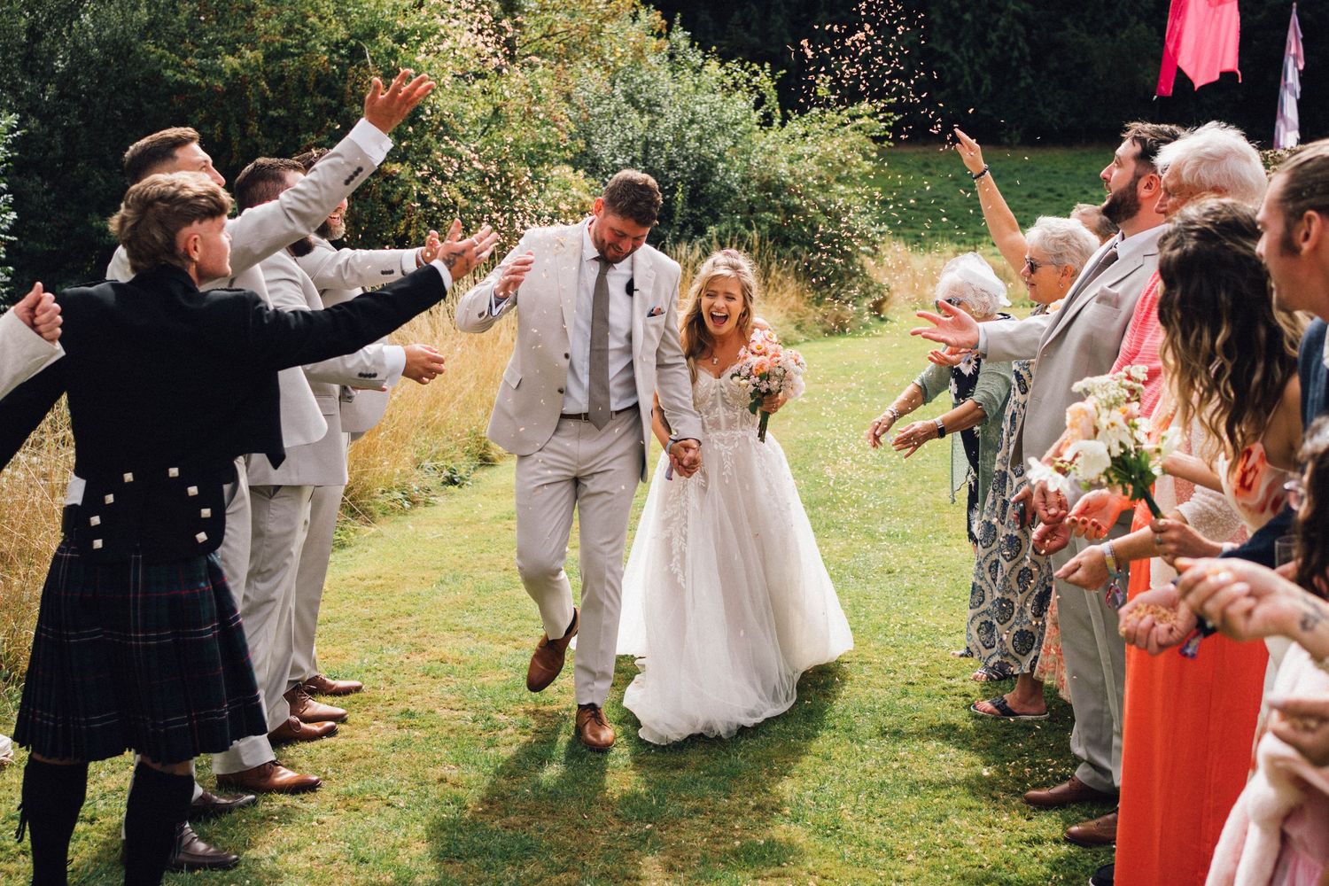 Wedding couple celebrates while walking through guests throwing confetti in an outdoor setting.