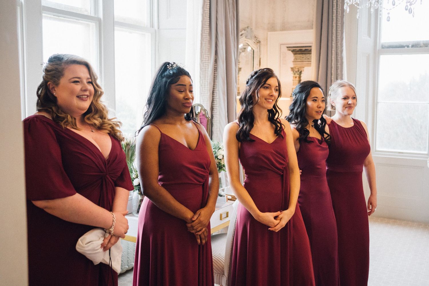 Bridesmaids in burgundy dresses laughing together in a sunlit room.