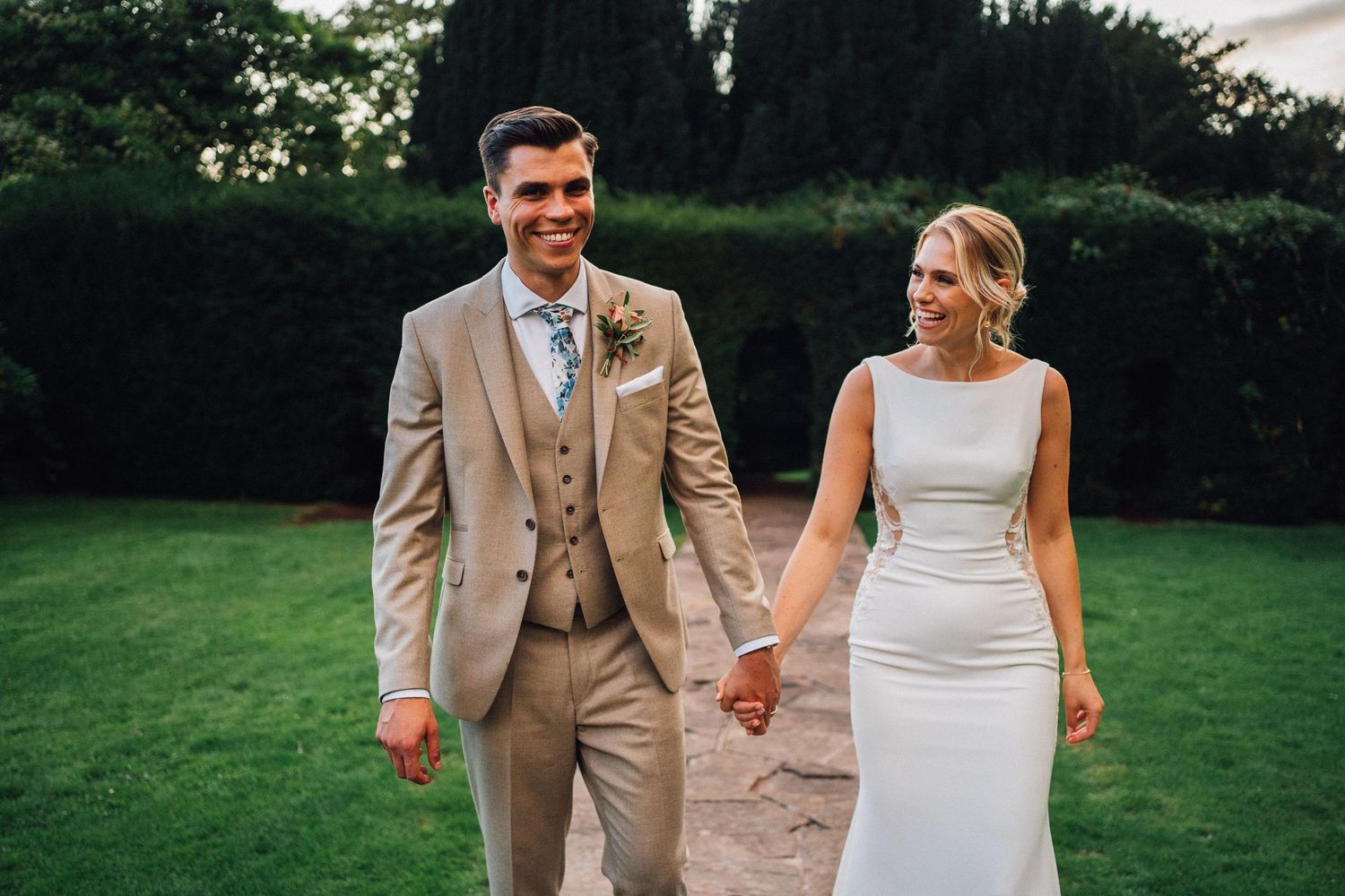 Wedding couple walking hand in hand across a green lawn during outdoor photography session.