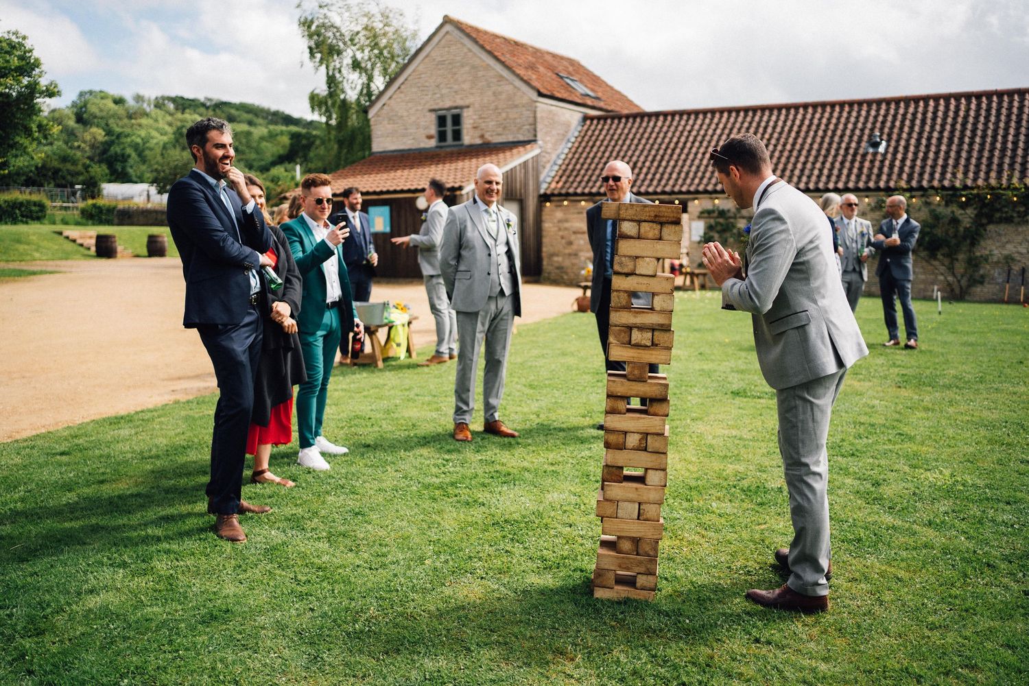 Wedding party playing oversized Jenga game on grass outside rustic venue.