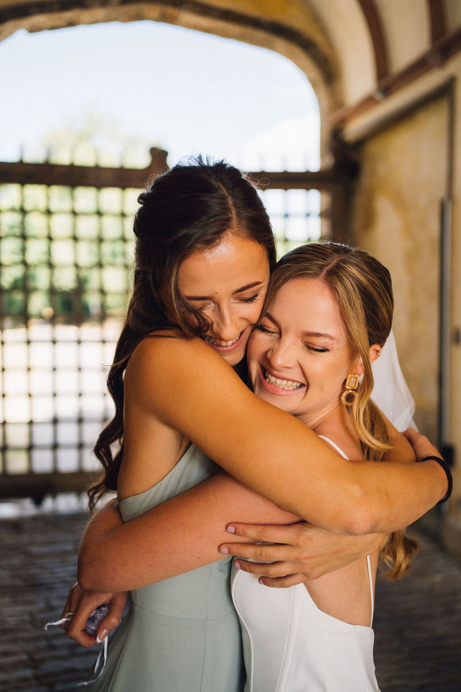 Two friends share a joyful embrace and laugh together while wearing formal dresses in an arched passageway.