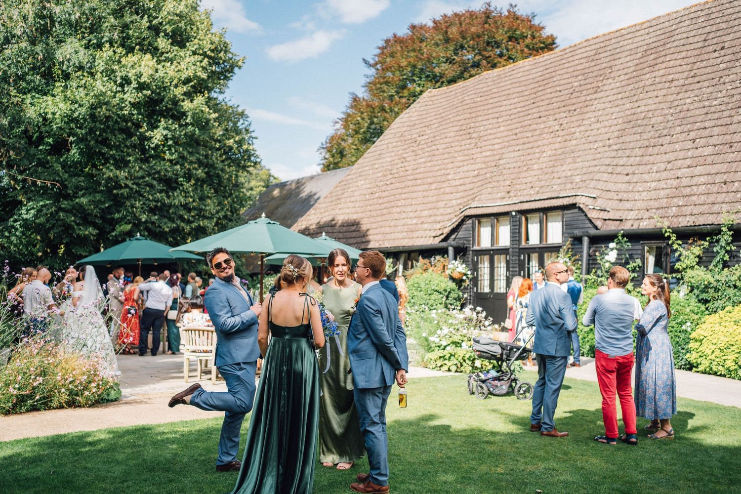 Outdoor wedding venue with guests mingling on a lawn surrounded by traditional buildings and green canopies.