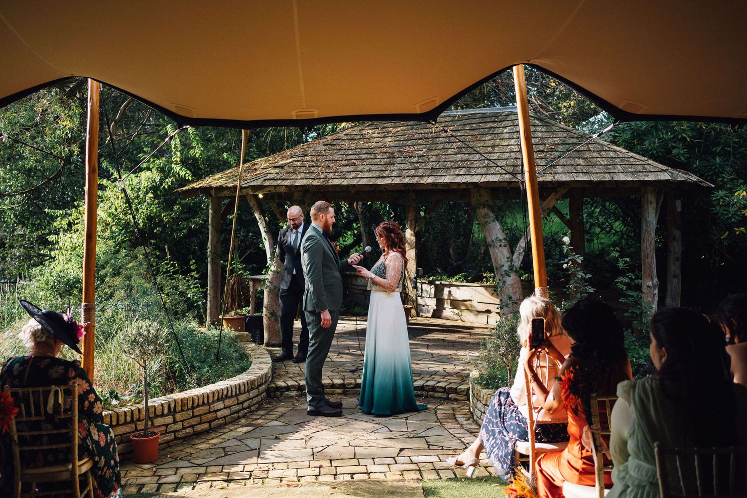 Outdoor wedding ceremony under a wooden gazebo with guests gathered in a lush garden setting.