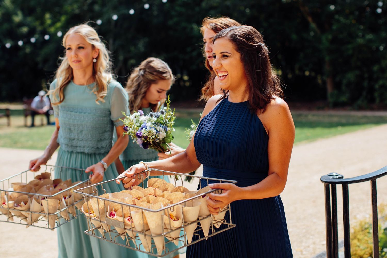 Bridesmaids in sage and navy dresses carry cone favors and bouquets at an elegant outdoor wedding.