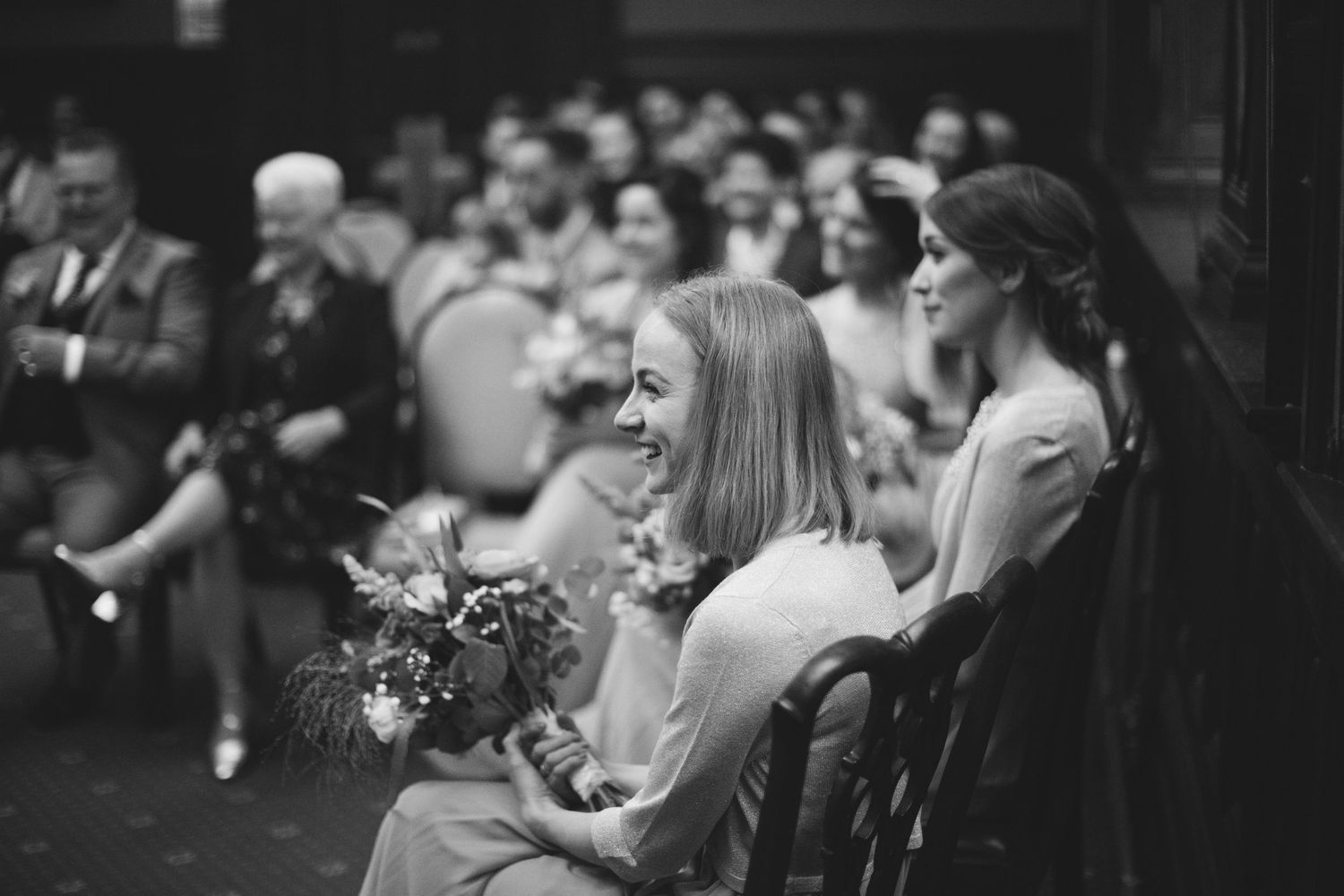 Black and white photo of smiling wedding guests seated, woman holding bouquet laughing joyfully during ceremony.