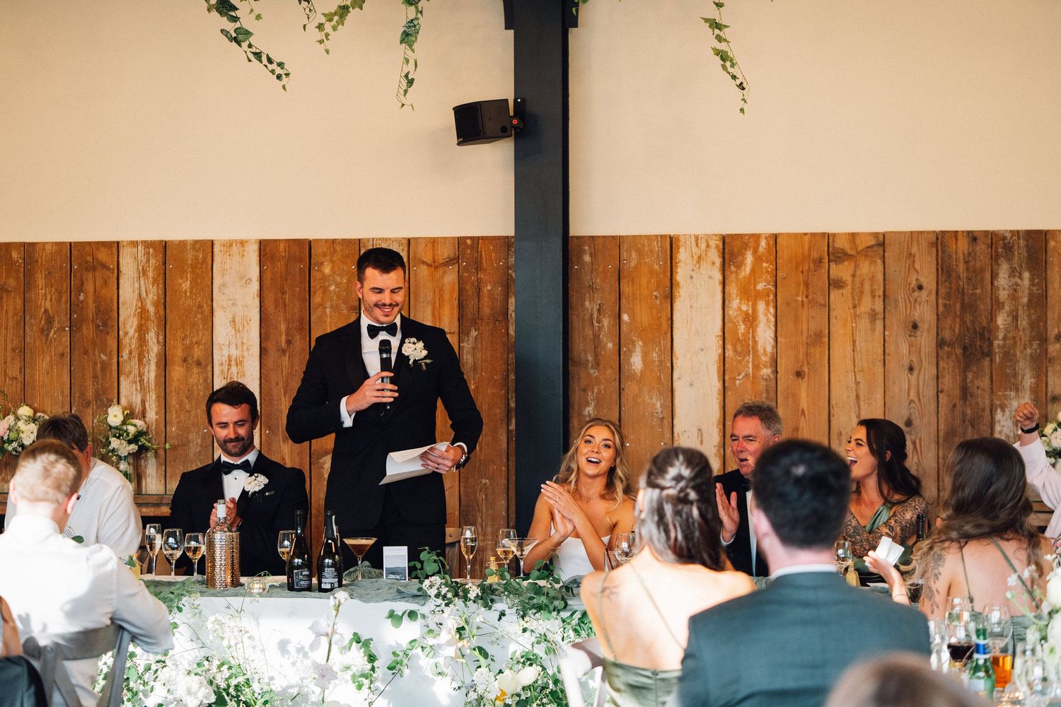 Wedding reception guests seated at long tables inside a rustic wooden barn venue with decorative lighting.