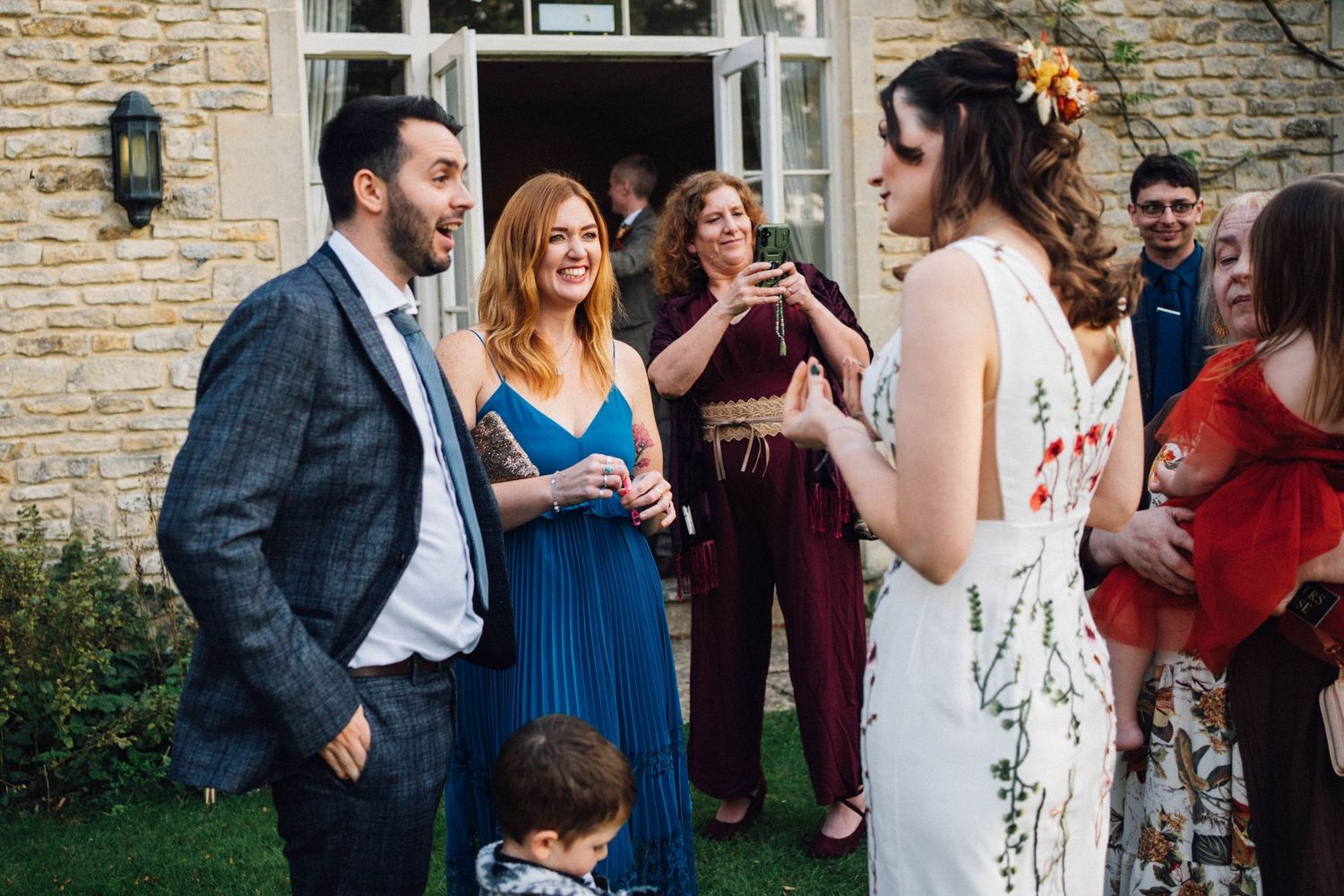Wedding guests laugh and socialize during an outdoor reception by a stone wall.