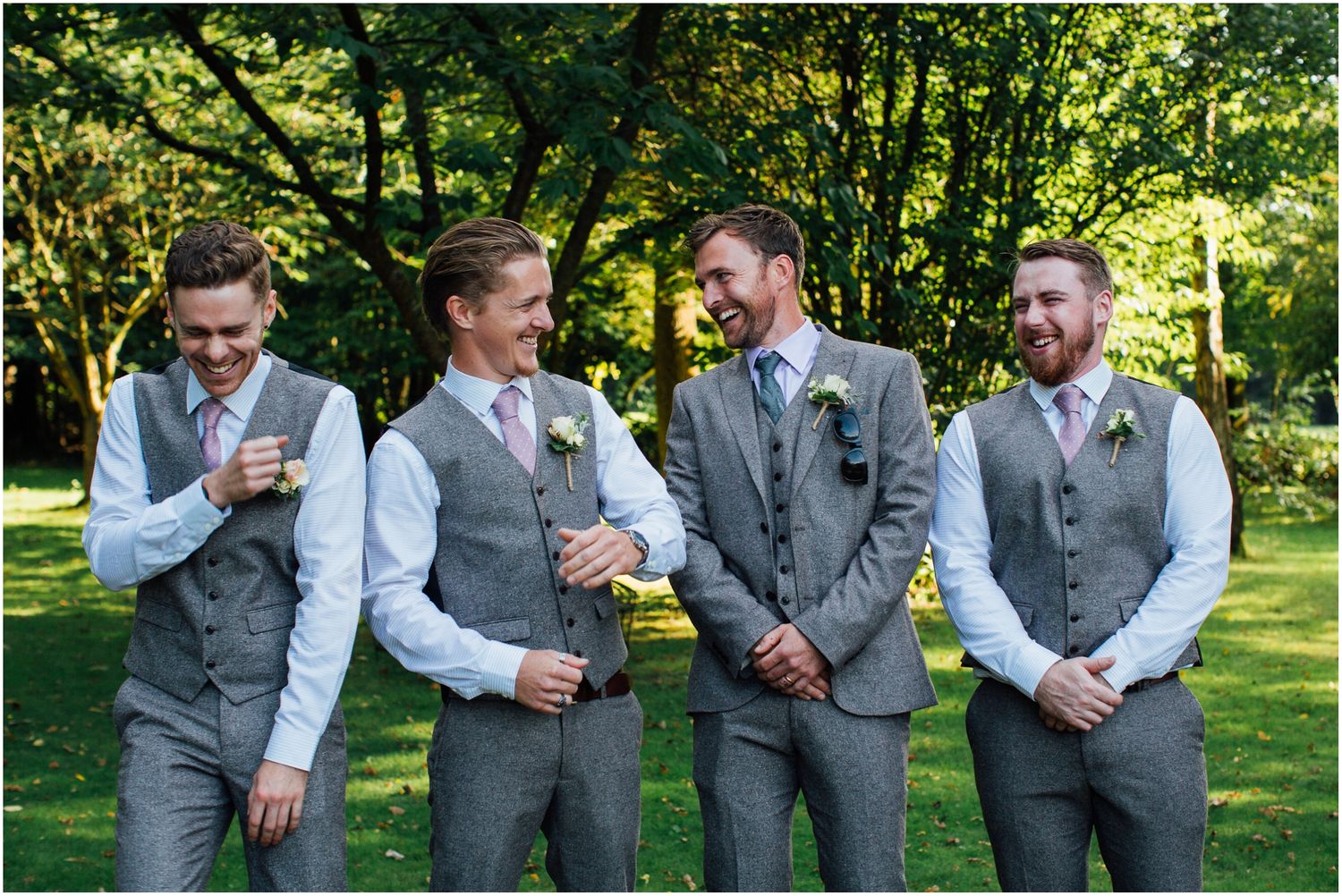 Wedding party groomsmen in grey suits share laughs outdoors.