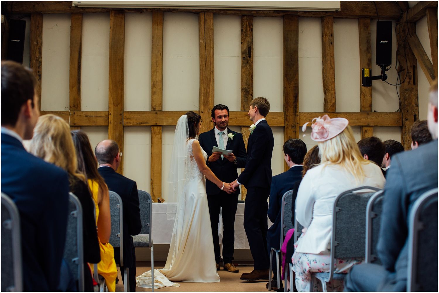 Wide view of wedding ceremony in progress inside timber-framed barn with white walls.