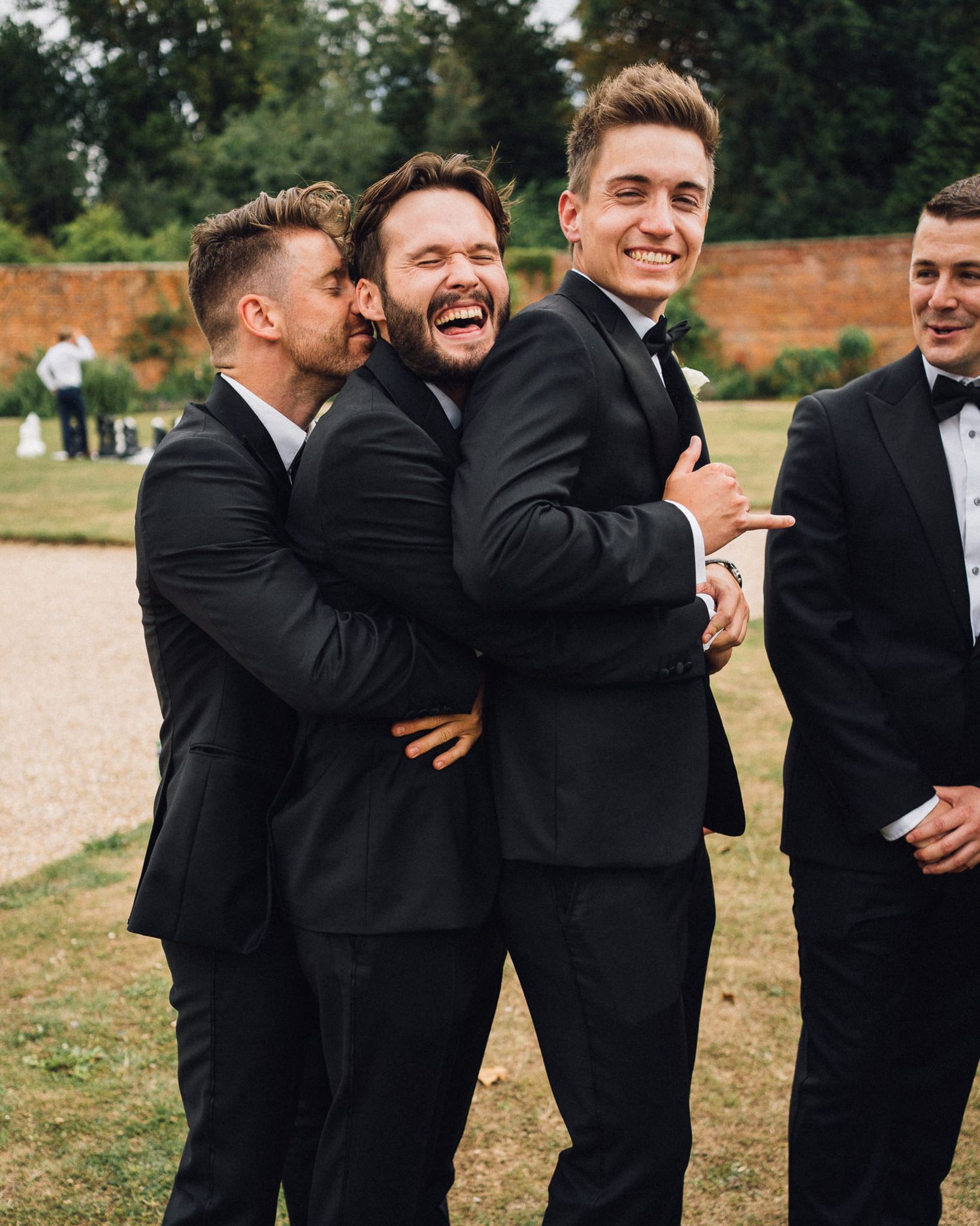 Groomsmen in black suits sharing laughter and embraces during outdoor wedding photo session.