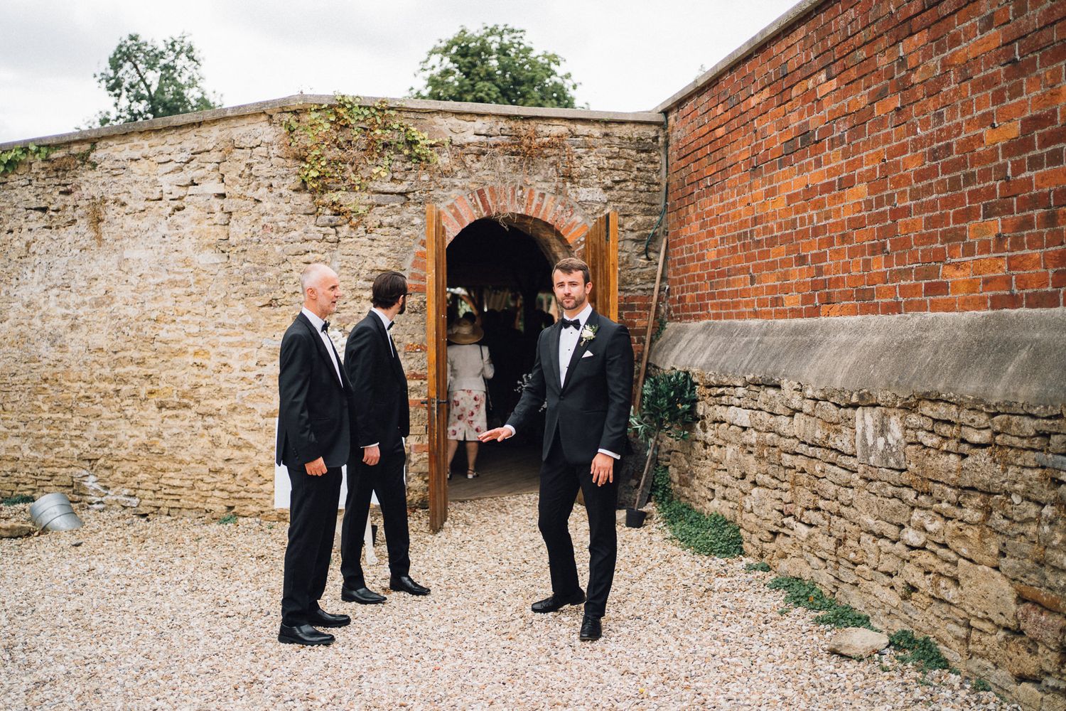 Groomsmen in black suits walk along a rustic brick wall pathway.