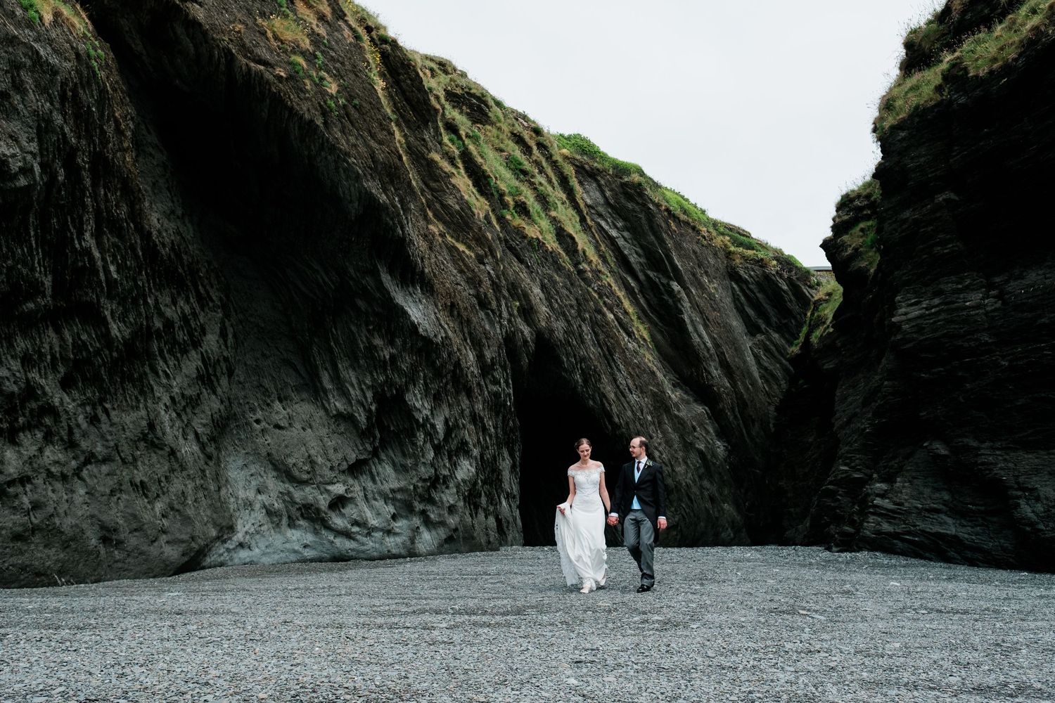 A couple walking on a black sand beach near dramatic rocky cliffs and a cave entrance.