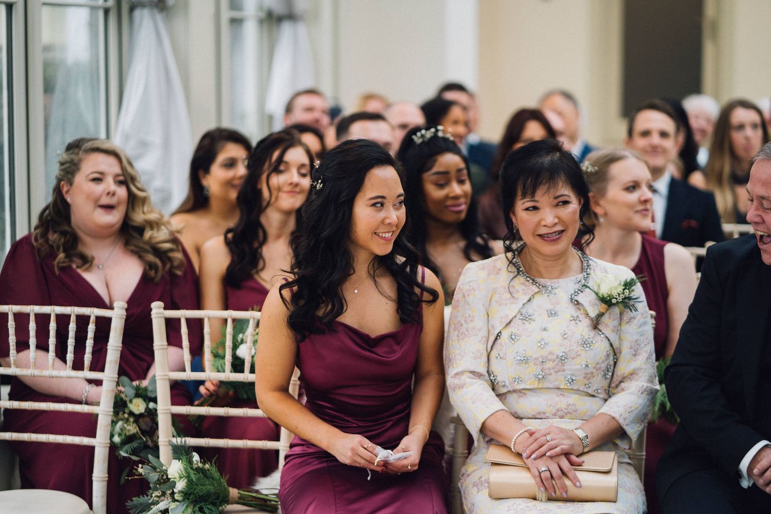 Wedding guests in burgundy dresses seated in rows during an indoor ceremony.