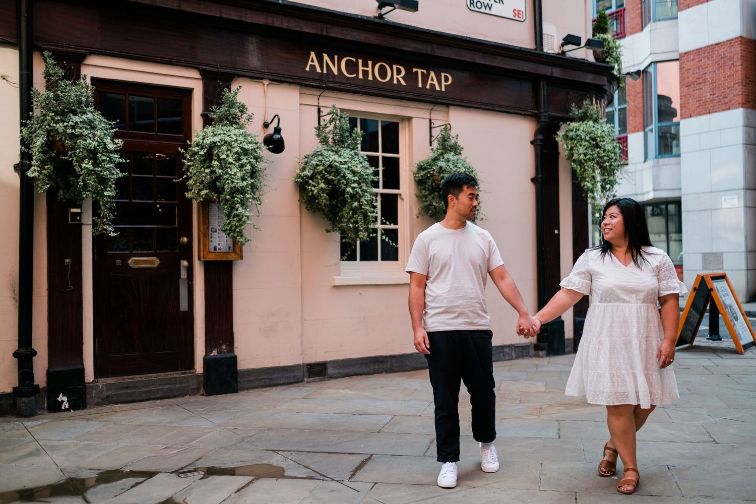 A couple walks together on a charming cobblestone street with pink buildings and hanging flower baskets.