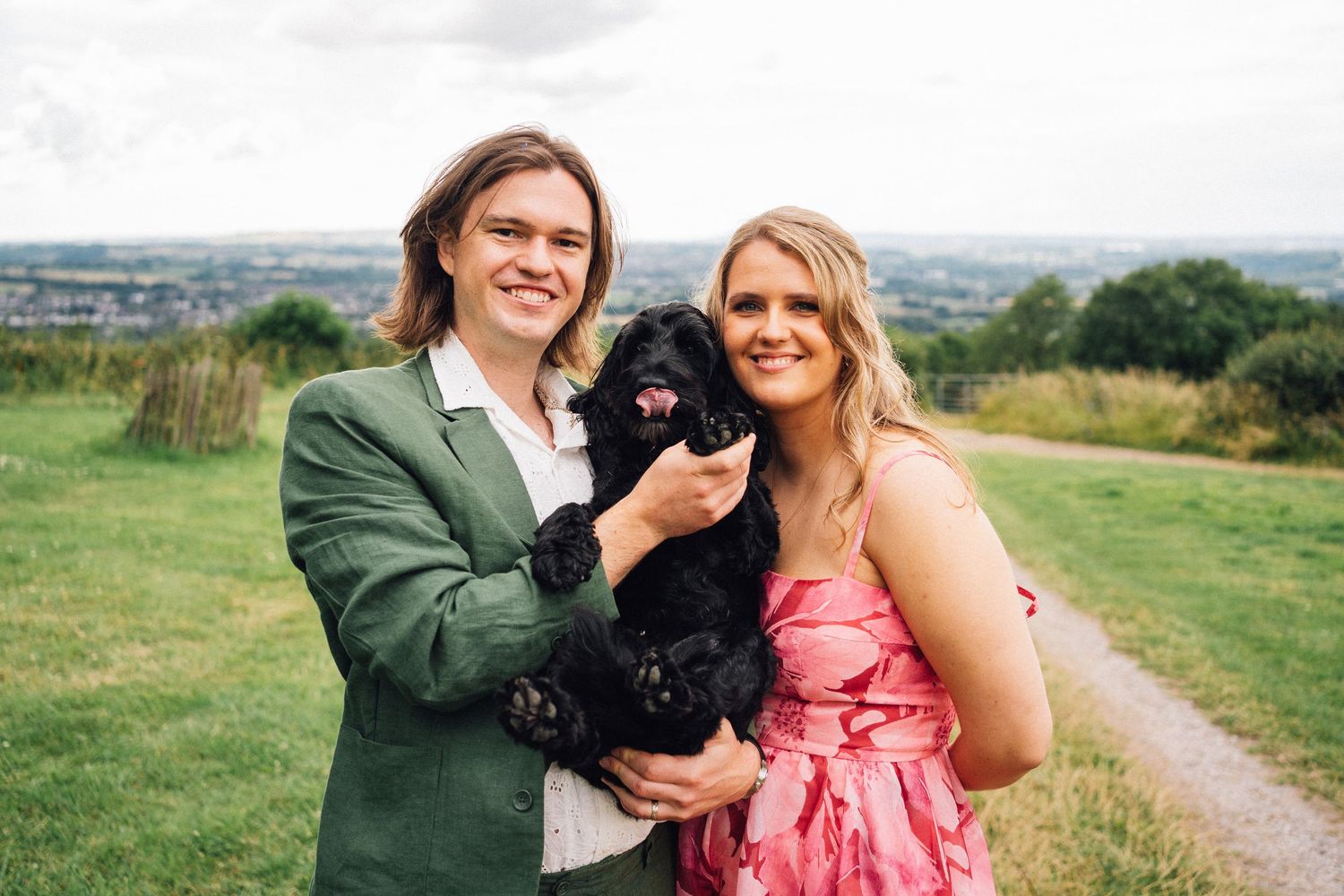 Wedding couple poses outdoors with black dogs in formal attire on a country path.