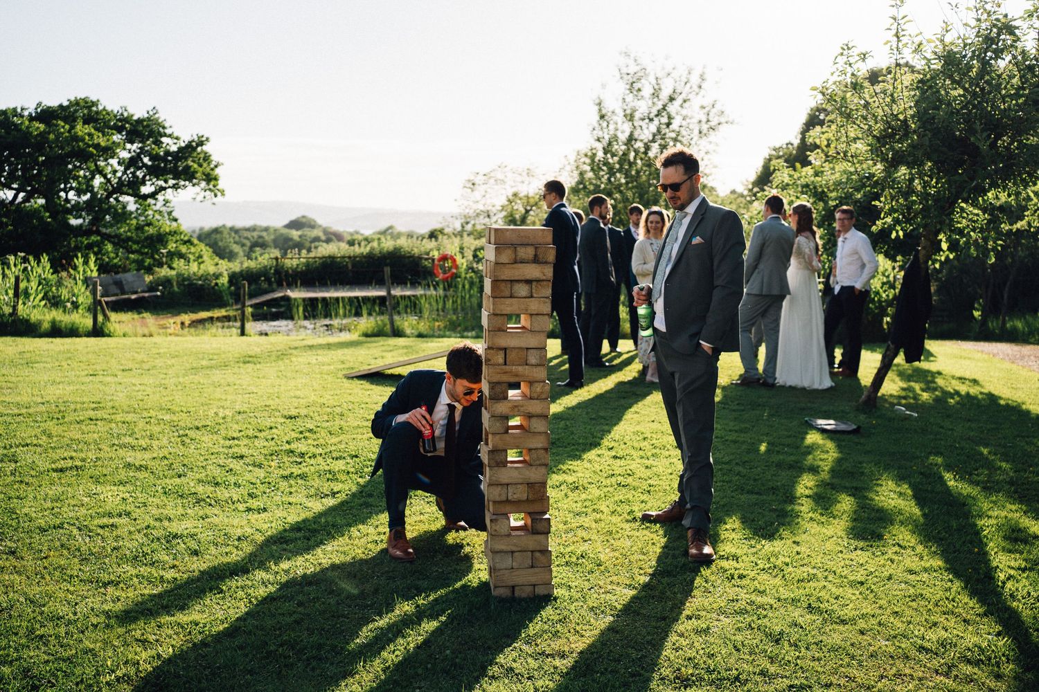 Wedding guests gather on a sunny lawn outside a rustic brick venue during golden hour.
