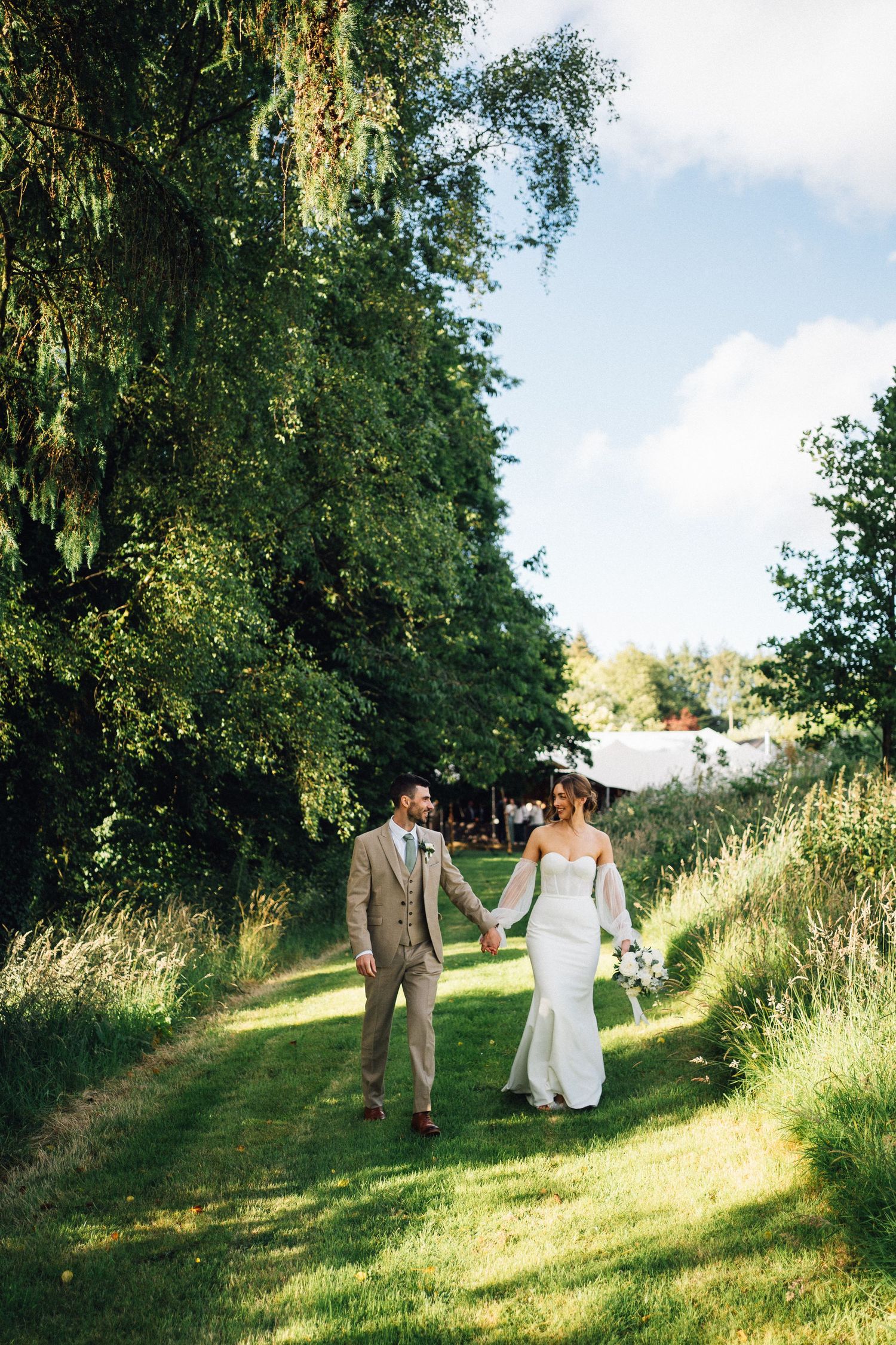 A couple walks hand in hand up a sunny hillside path surrounded by lush summer greenery.