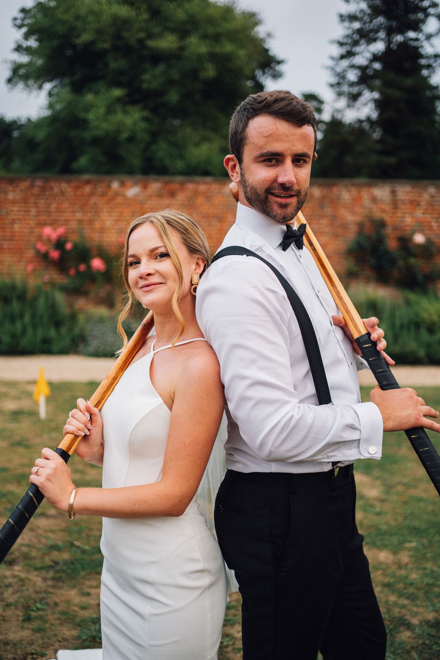 A couple posing playfully with golf clubs in a garden setting on their wedding day.