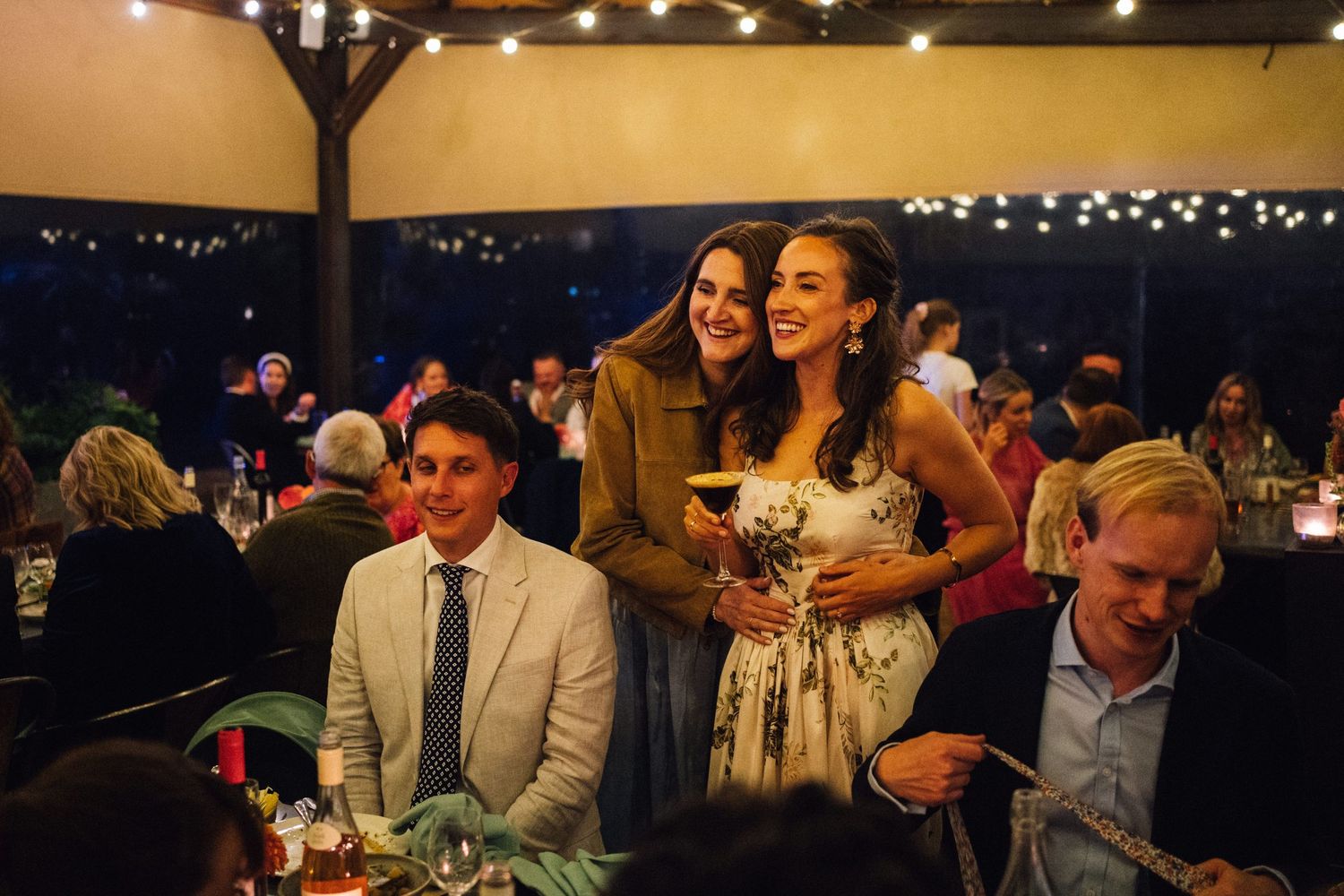 Wedding guests enjoying a lively evening reception with string lights and warm ambiance at an indoor venue.