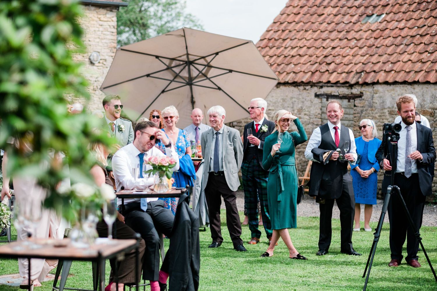 Wedding party and guests mingle on a garden lawn with umbrellas during an outdoor reception.