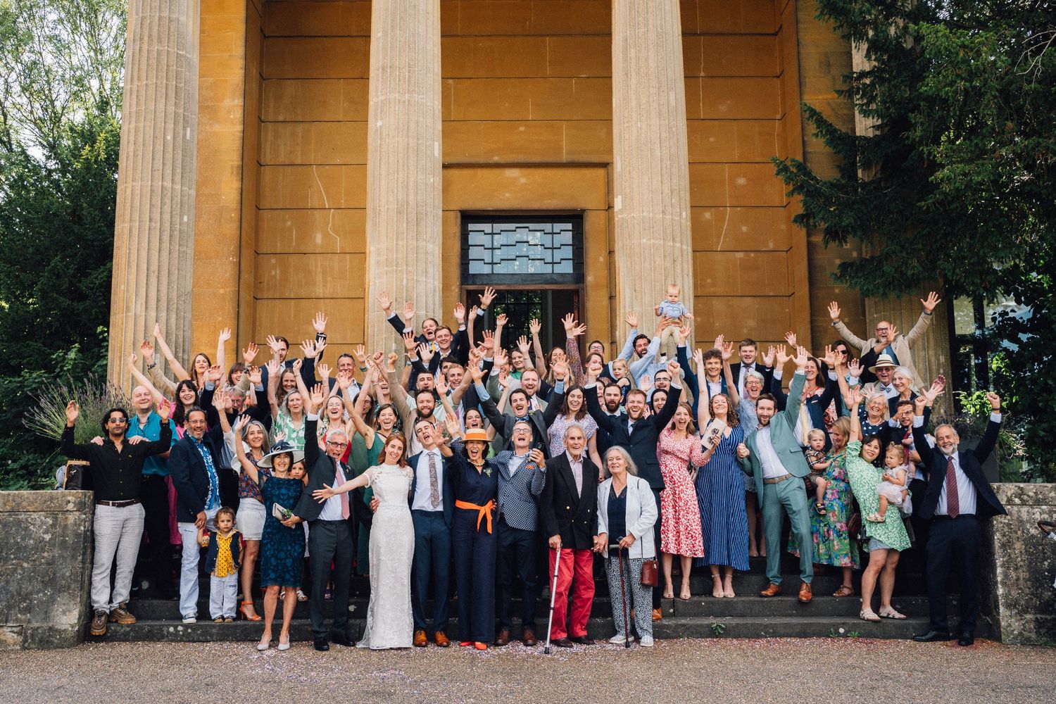 Large wedding party poses for group photos outside a classical stone building with columns.