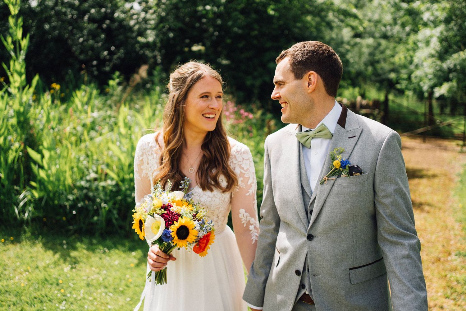 A couple in wedding attire share a joyful moment together outdoors in a sunny garden setting.
