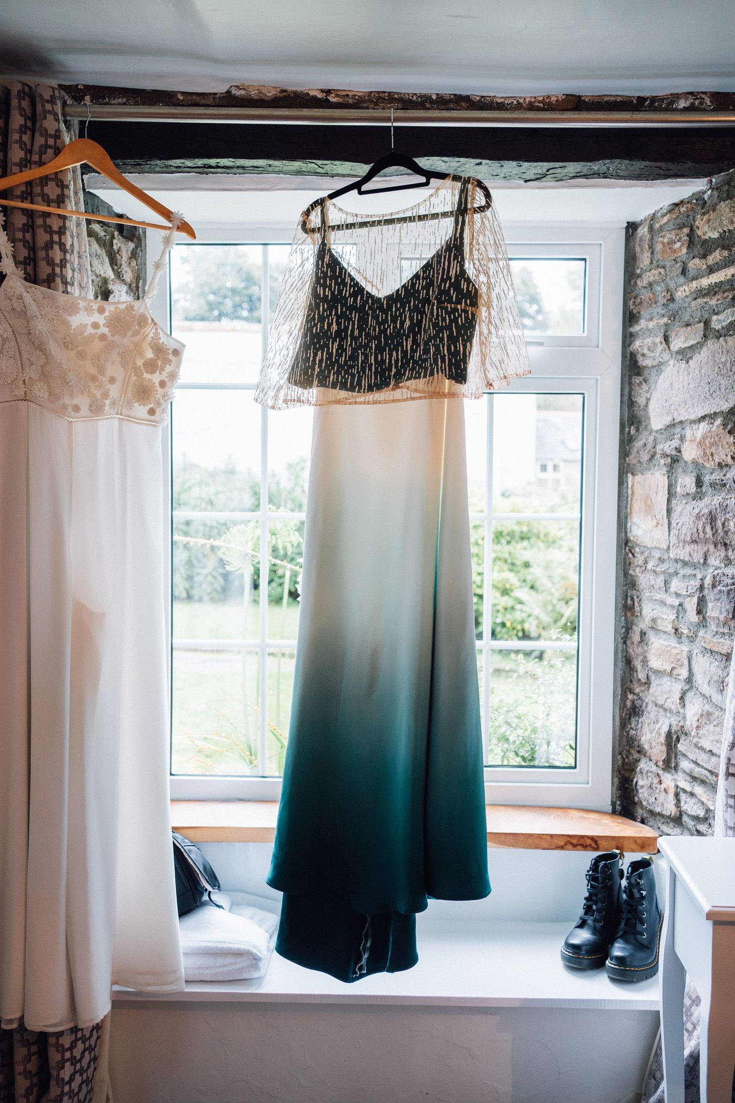 Wedding attire including dresses and suits hanging by windows in a rustic stone-walled room with natural lighting.