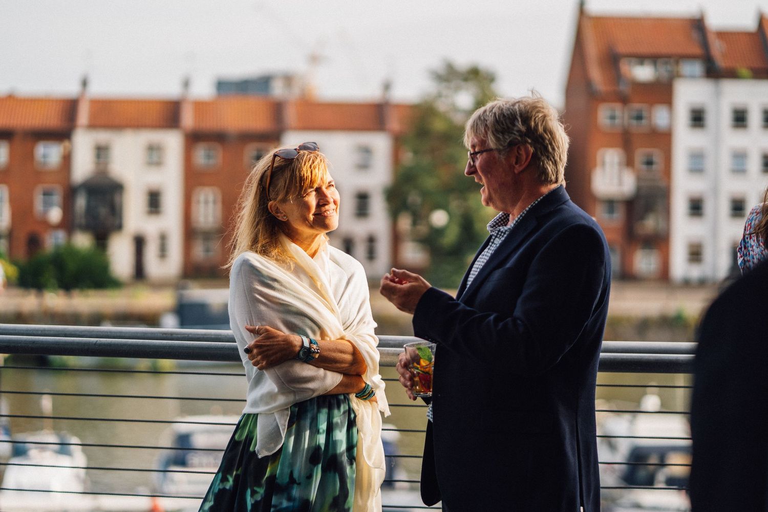 Two people conversing on a riverside balcony with red brick buildings in the background.
