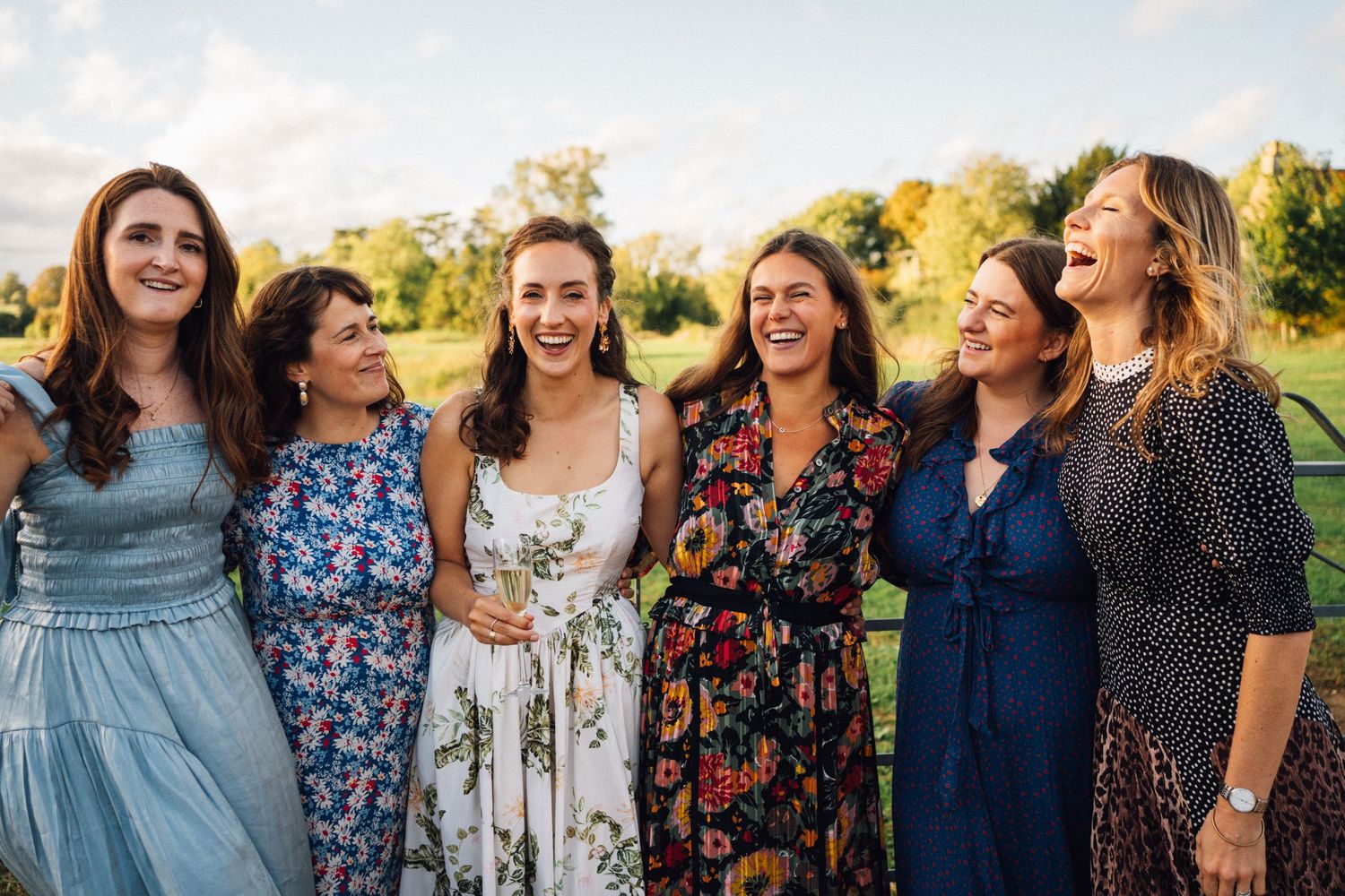 A group of women in floral dresses and casual attire laugh together outdoors during a sunny afternoon gathering.