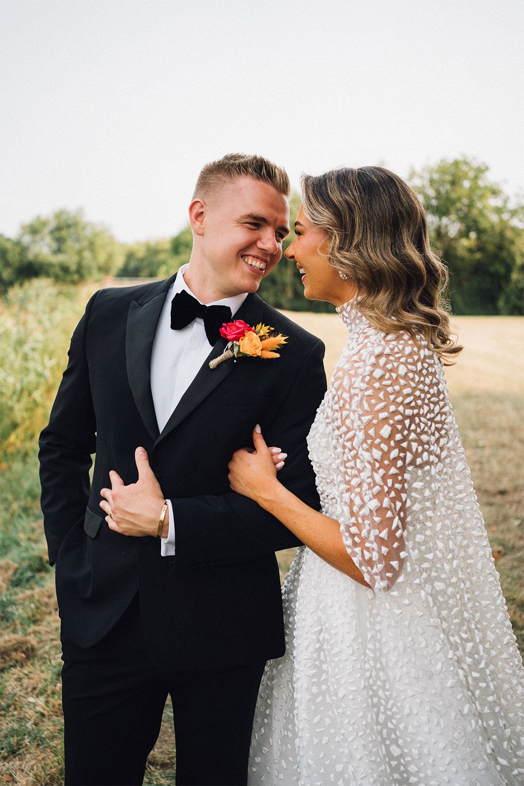 A couple in wedding attire share an intimate moment outdoors, with the bride in a lace gown and groom in black tuxedo.