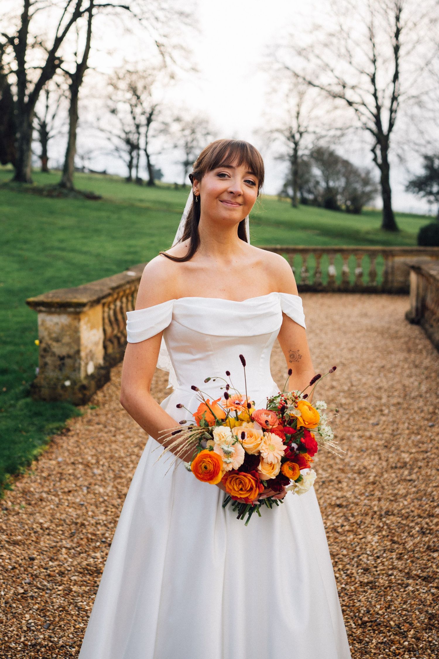 A bride in an off-shoulder white gown holds a vibrant orange and burgundy fall bouquet in an outdoor autumn setting.