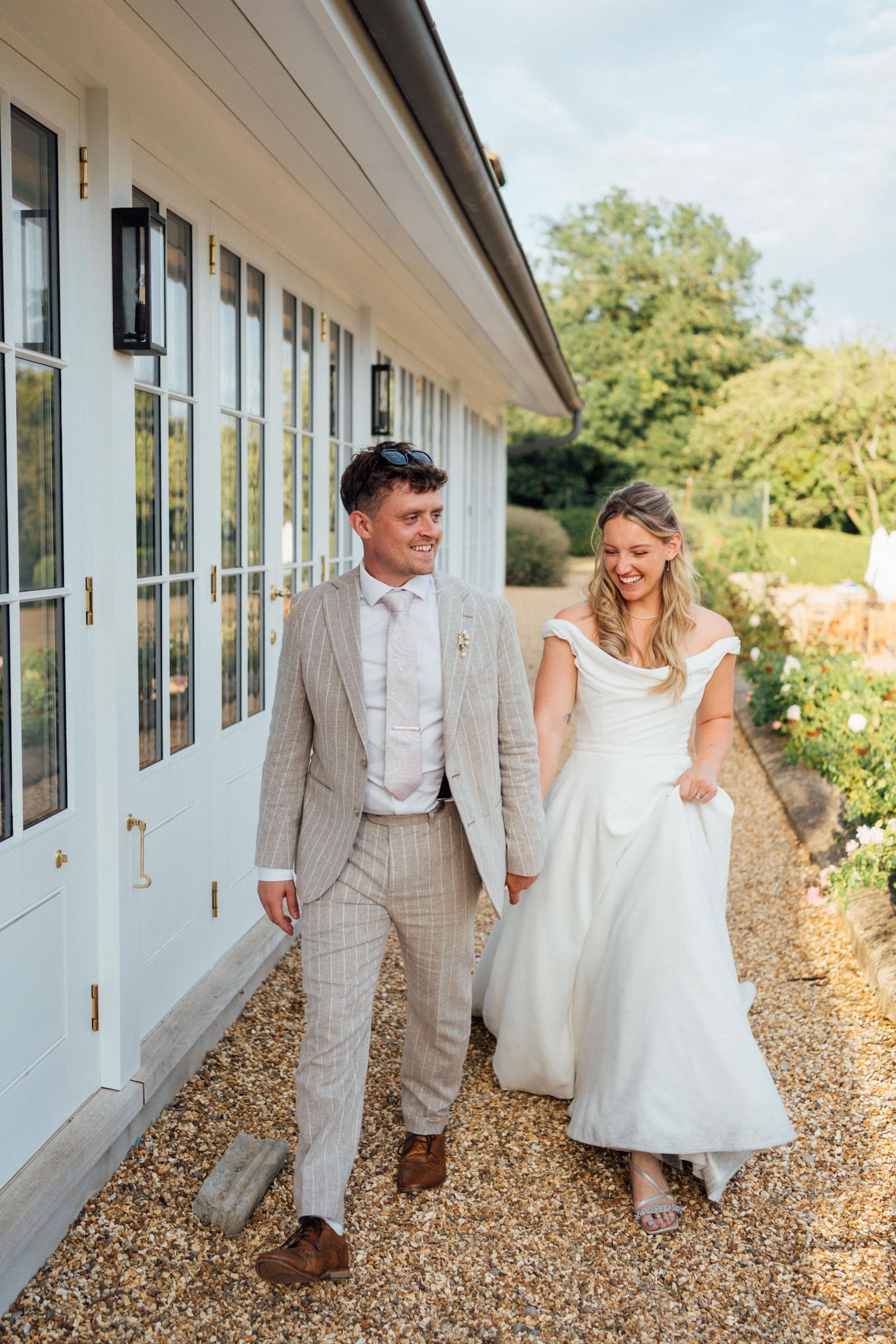 A smiling bride and groom walk hand in hand outside a white country venue on their wedding day.
