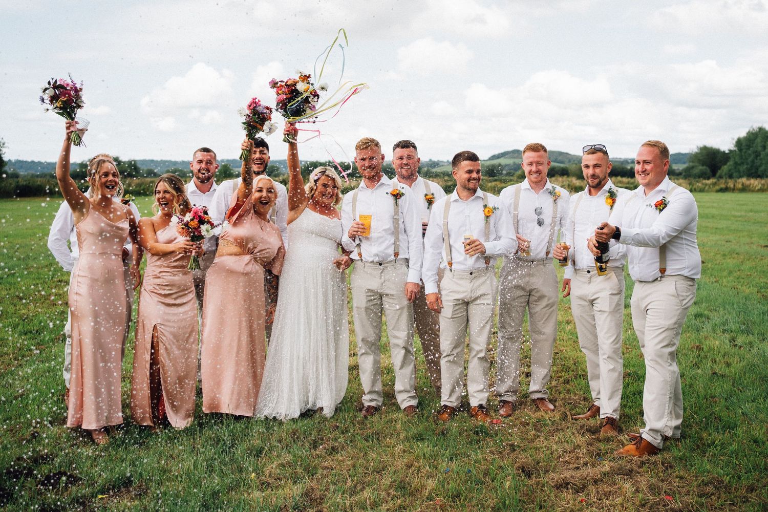 Wedding party in blush pink dresses and beige suits poses together in a sunny outdoor field.