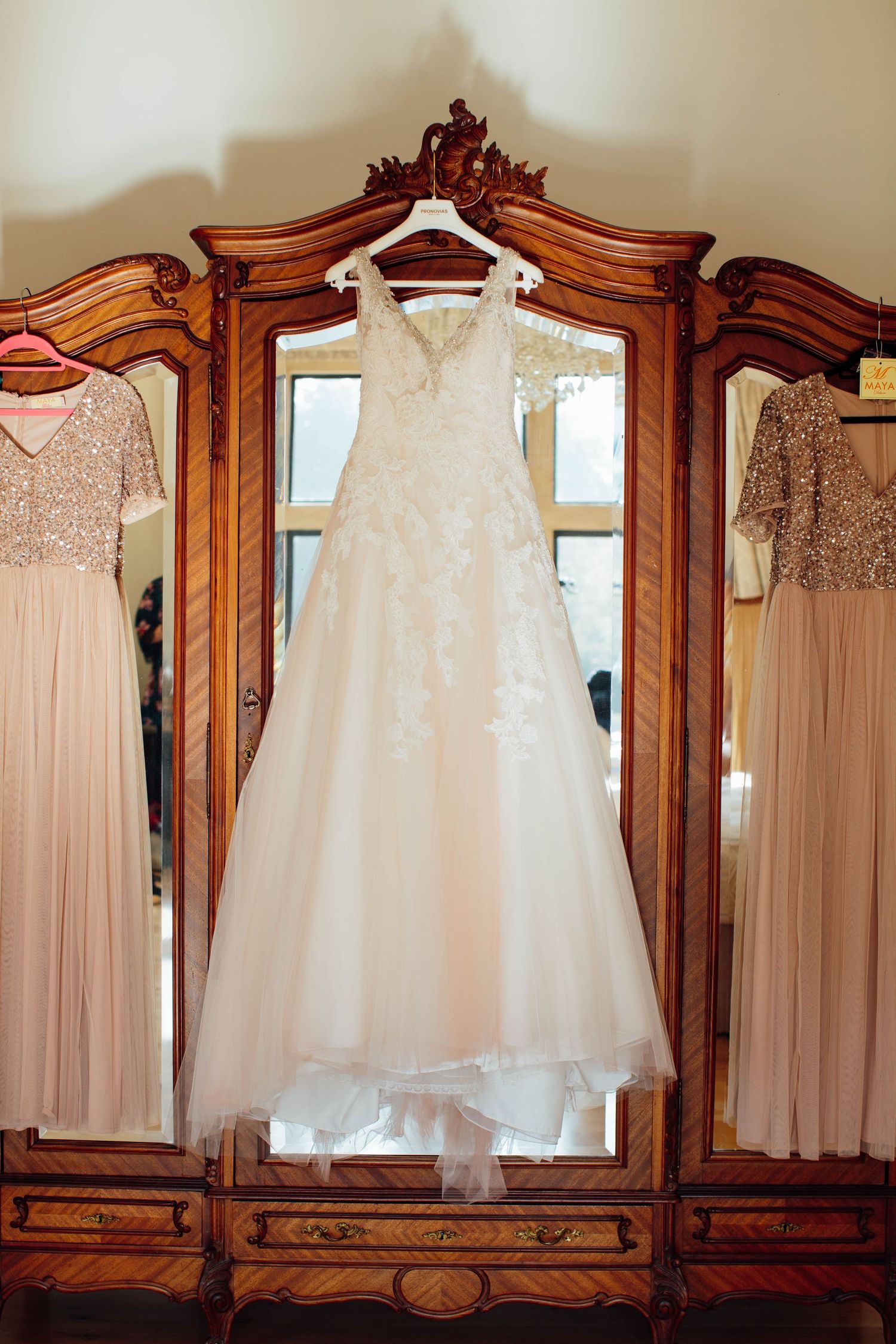 Elegant white lace wedding dress hanging on ornate vintage wooden armoire with blush bridesmaid dresses on sides.