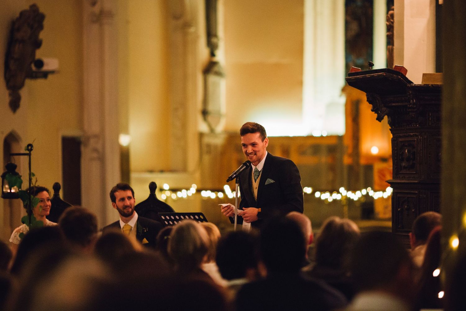 A wedding speech being delivered in a warmly lit church setting with string lights.