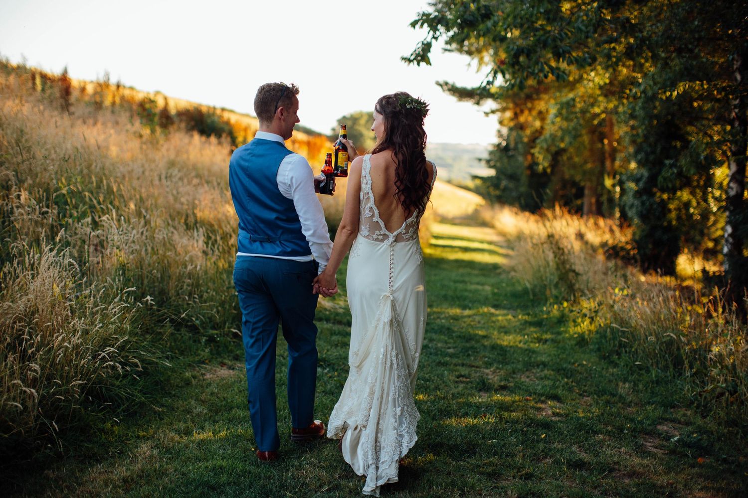 Newlyweds walking hand in hand through golden field at sunset.