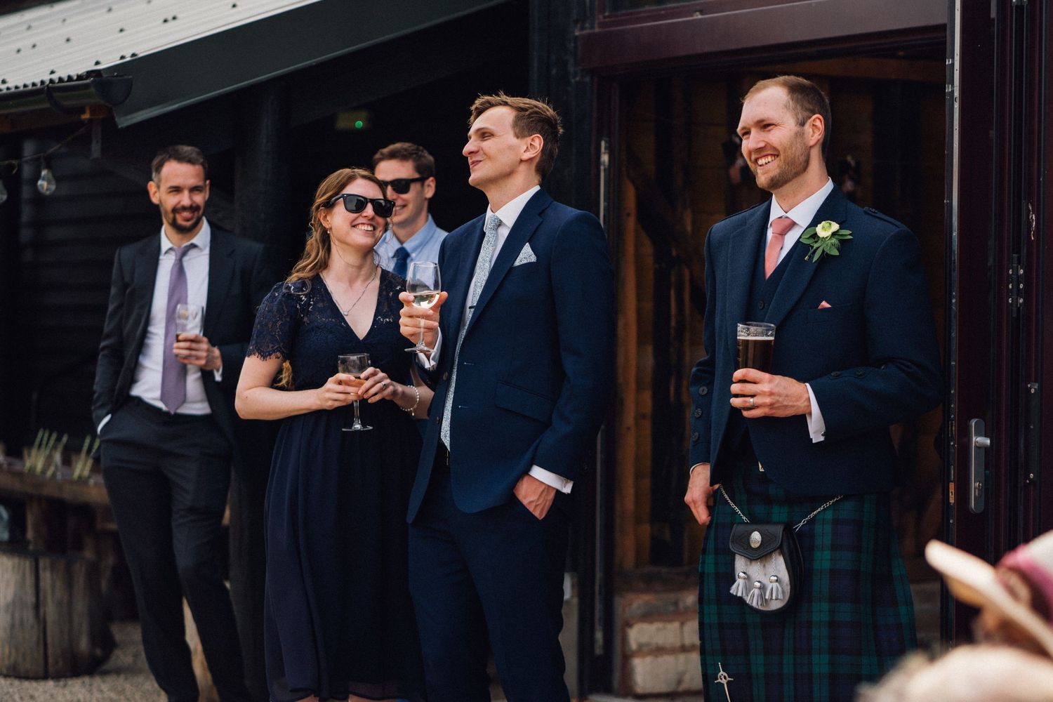 Wedding guests in formal attire and traditional Scottish kilts enjoying drinks at an outdoor reception.