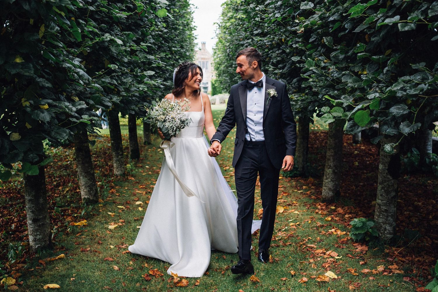 Wedding couple walking through a tree-lined path with autumn leaves on the ground.