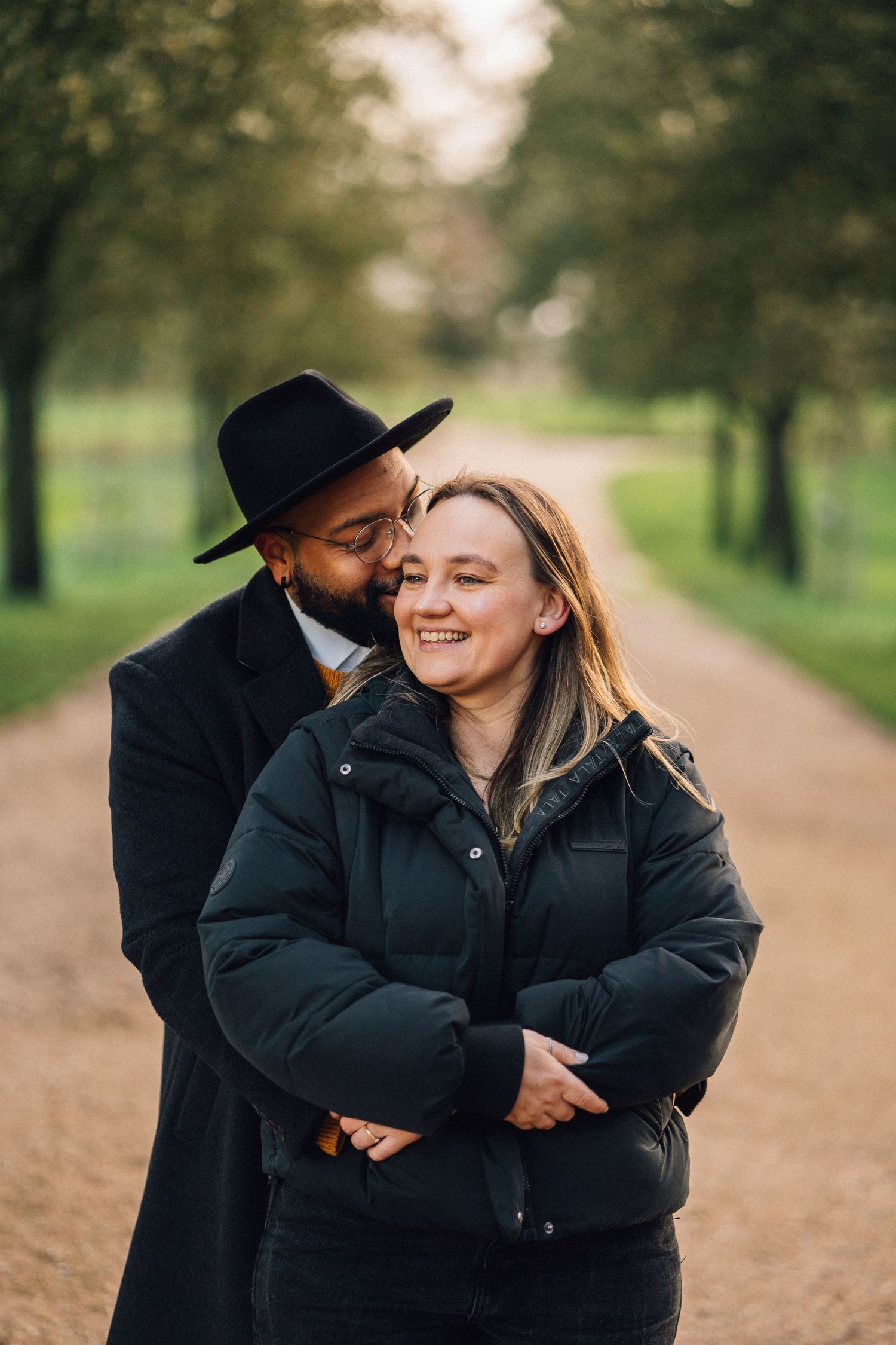 A romantic couple embraces and laughs together on a scenic path surrounded by trees.