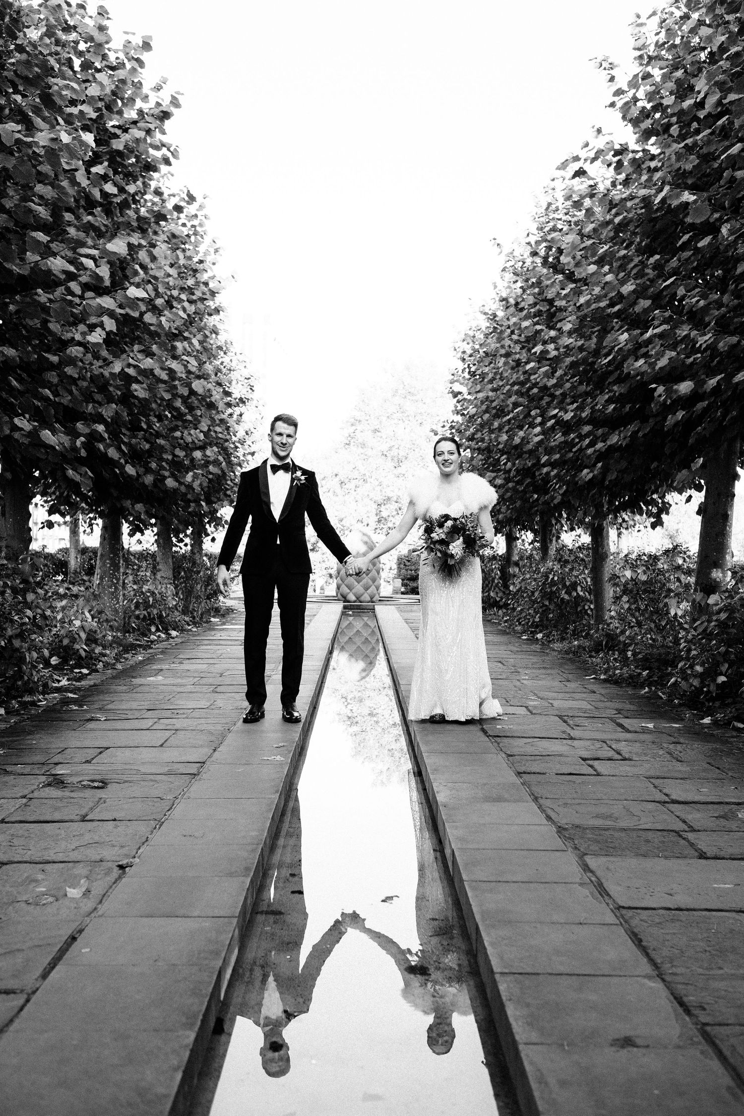 A black and white wedding photo with a reflection in a water feature between tree-lined paths.