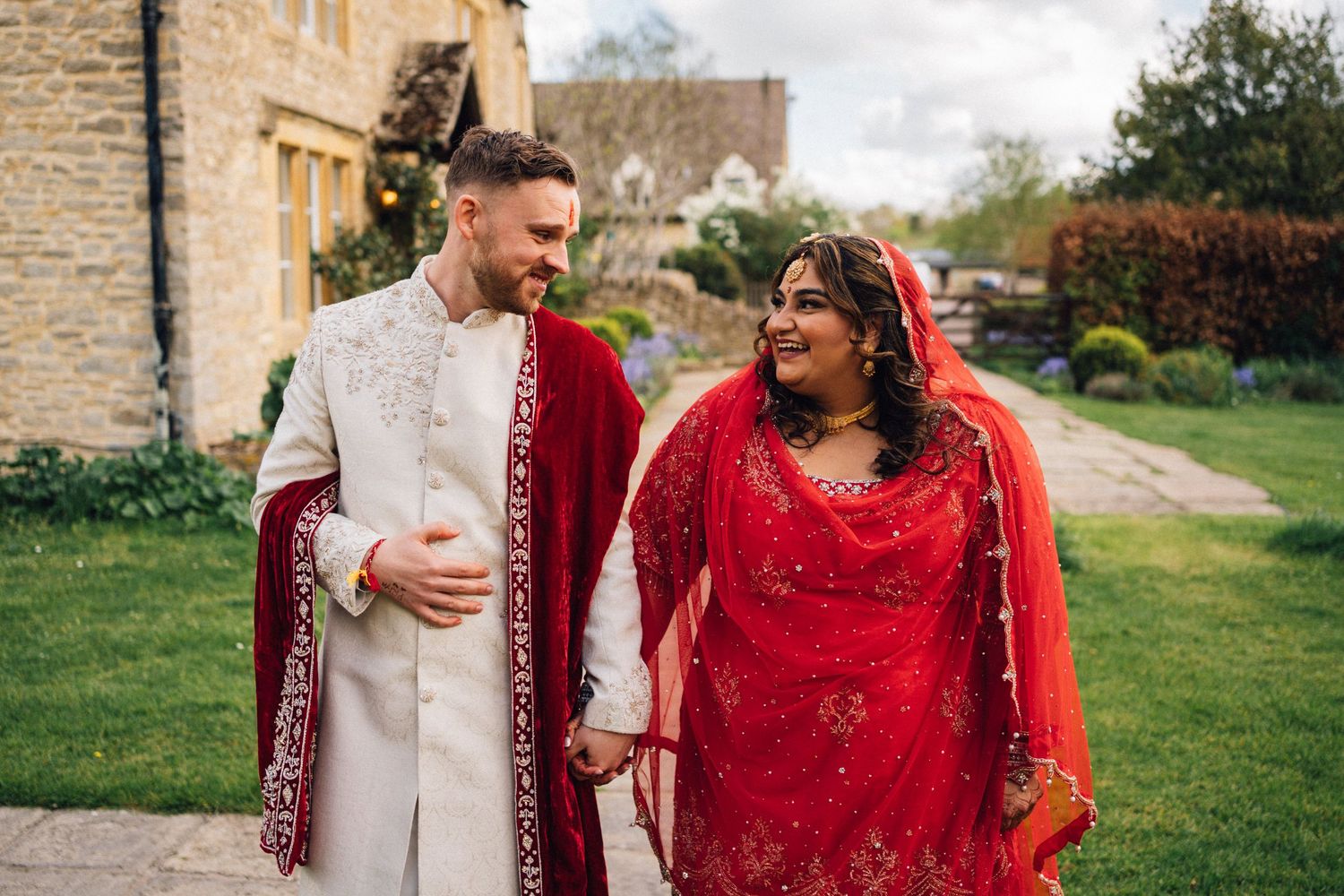 Couple in traditional South Asian wedding attire walking hand in hand in garden setting.