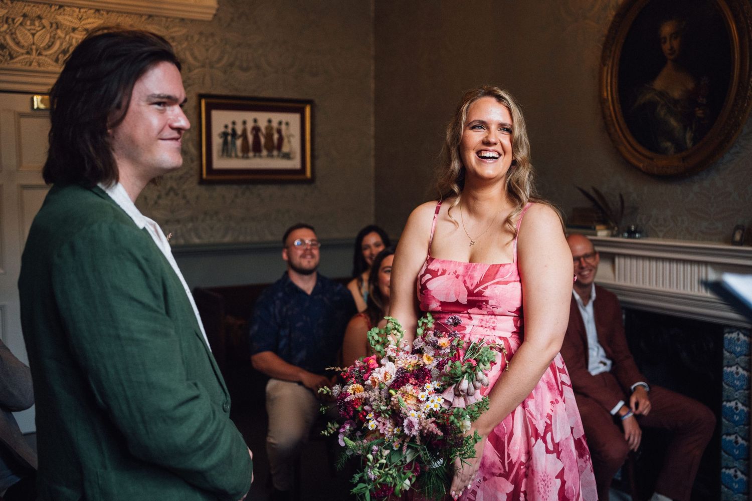 Bride in pink floral dress laughing with groom in green suit during intimate wedding ceremony indoors.
