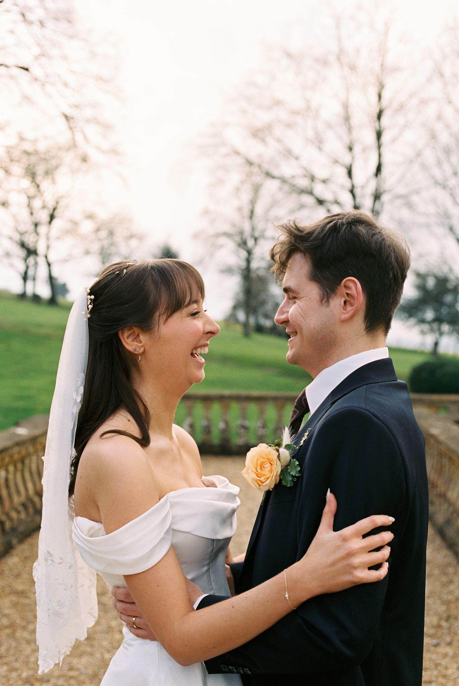 Bride and groom laughing together outdoors at a wedding, bride wearing off-shoulder gown with veil, groom in navy suit.